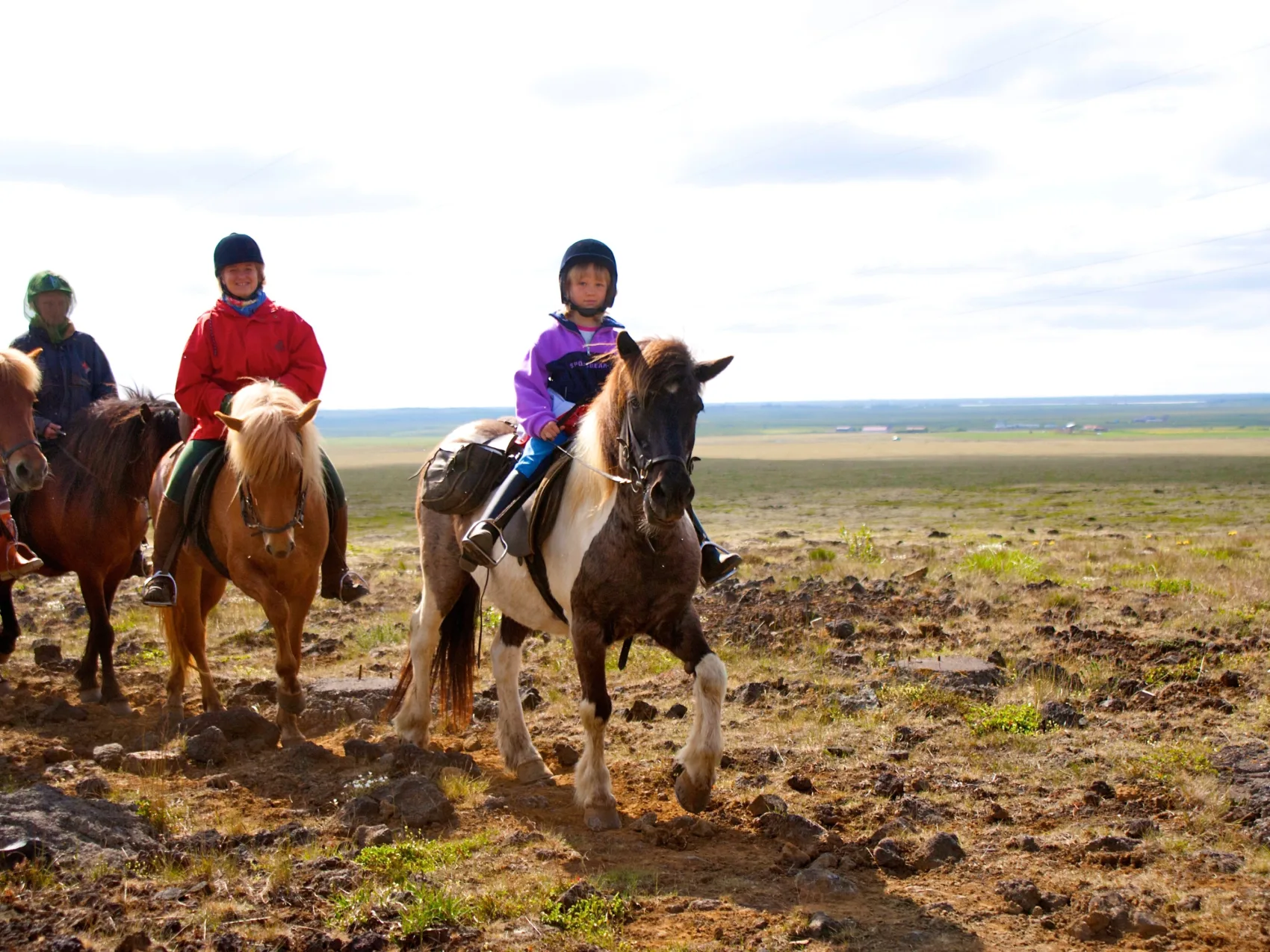 Reiten Island mit Eldhestar; 
Gruppe reitet über eine Ebene, die nur spärlich mit Gras bewachsen ist. Ein Kind auf einem gescheckten Islandpferd reitet voraus in der Mitte des Bildes.