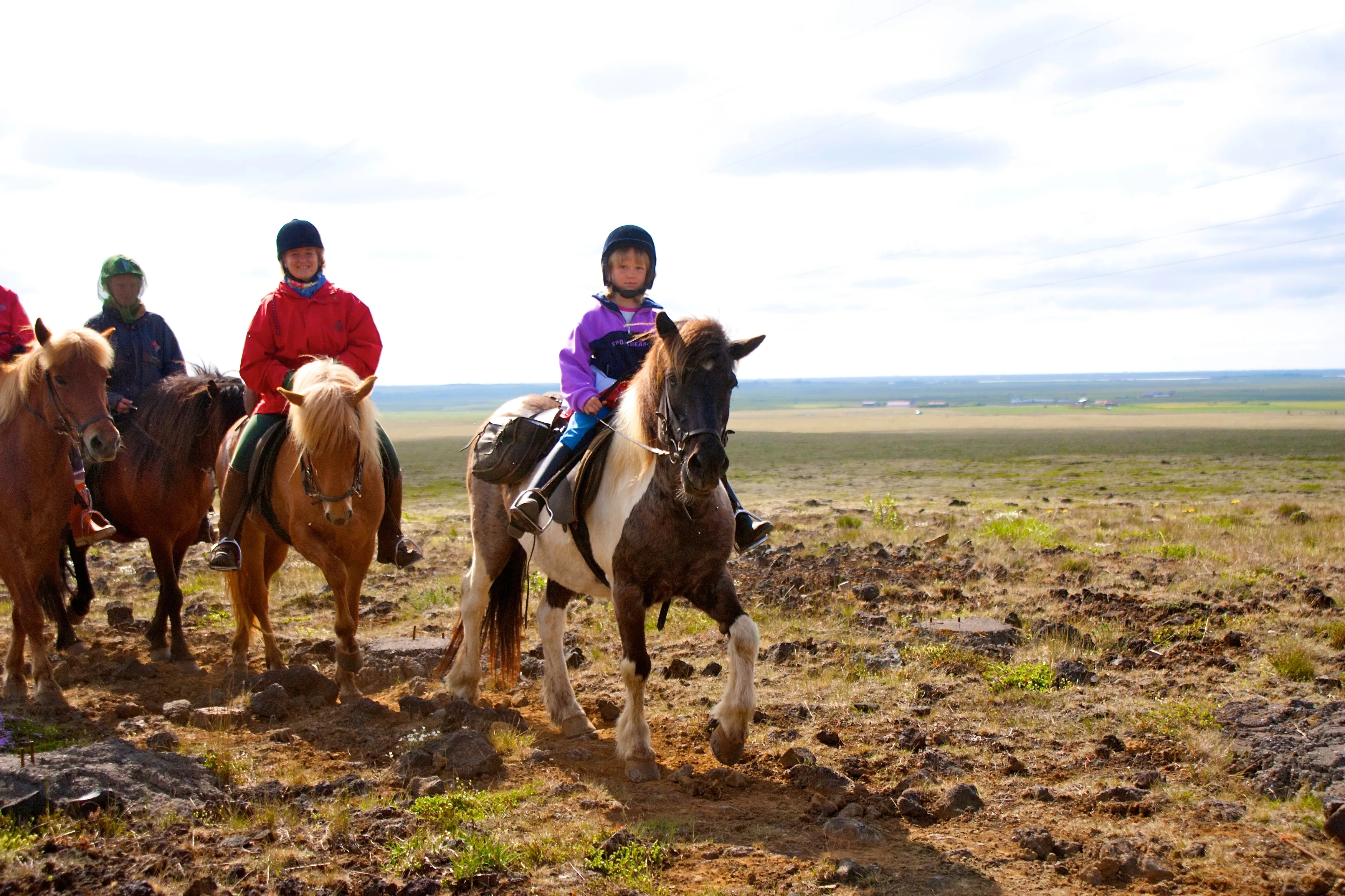 Reiten Island mit Eldhestar; 
Gruppe reitet über eine Ebene, die nur spärlich mit Gras bewachsen ist. Ein Kind auf einem gescheckten Islandpferd reitet voraus in der Mitte des Bildes.