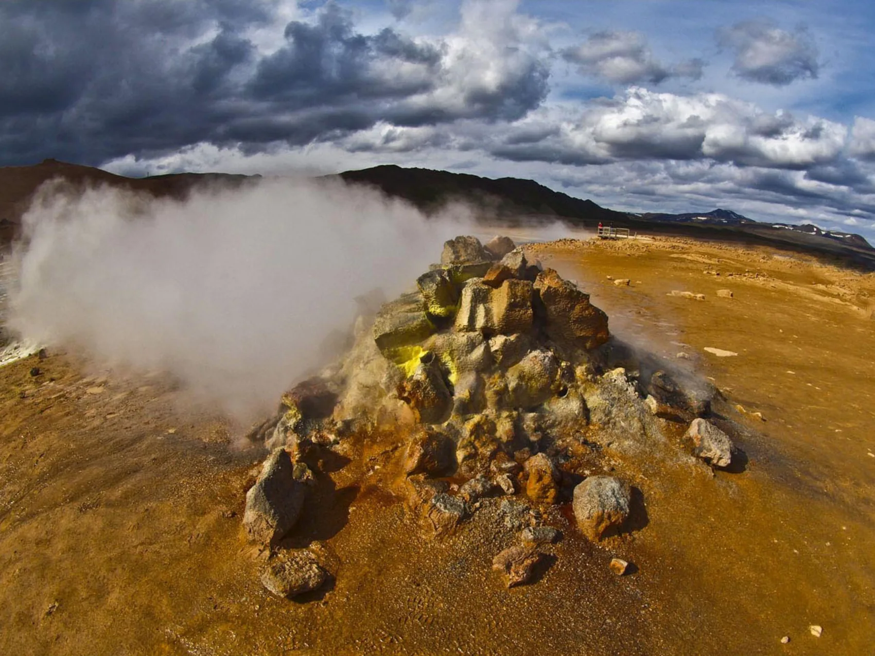 Hochtemperaturgebiet Hverarönd, Foto: Kurt Schneider
zeigt einen Steinhaufen, aus dem Dampf und Schwefel austreten, der Boden ist rot-gelb gefärbt. Im Hintergrund Wolken und Berge.