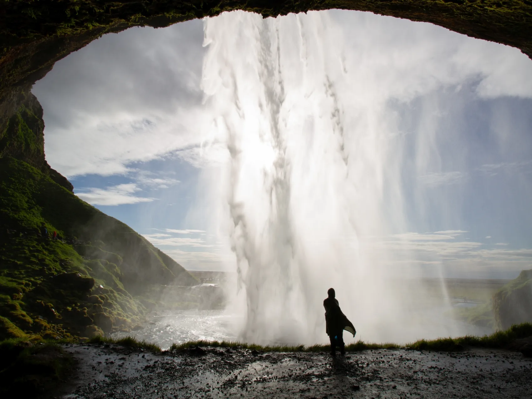 Person im Regencap steht hinter dem Wasserfall, den von vorne die Sonne anleuchtet