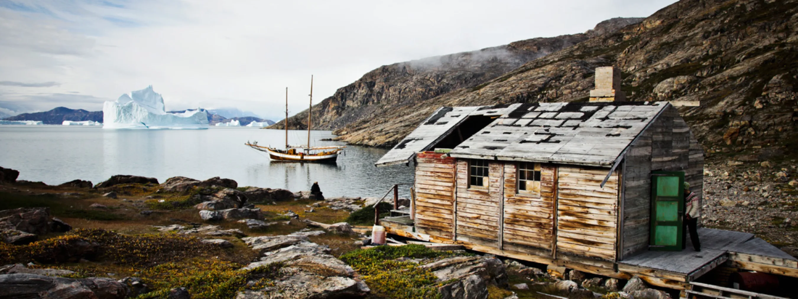alte, zerfallene Hütte, dessen Dach größere Löcher hat. Segelschiff im Fjord dahinter und Eisberge im Hintergrund