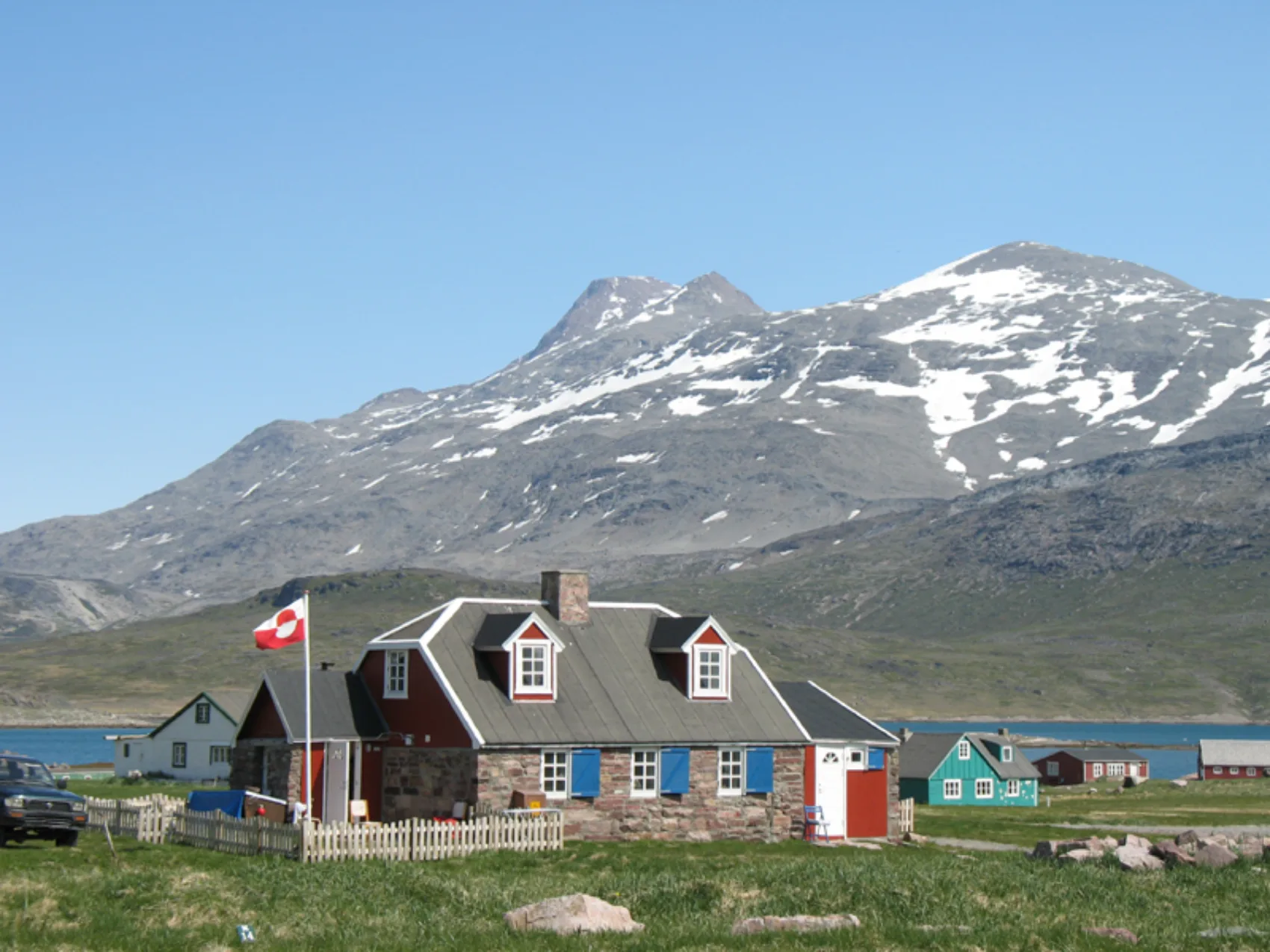 Steinhaus mit Grönland Fahne im Garten und leicht mit Schnee bedeckte Berge im Hintergrund