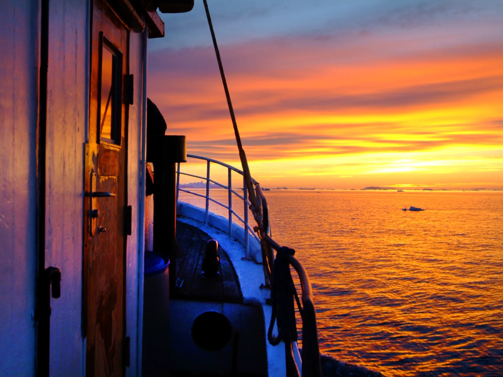 Ostgrönland Sonnenuntergang auf dem Boot, vereinzelte Eisberge schwimmen im Meer vor dem Bot, an dem seitlich vorbeigeschaut wird. Der Himmel leuchtet in Orange und Gelben Farben mit Wolken.