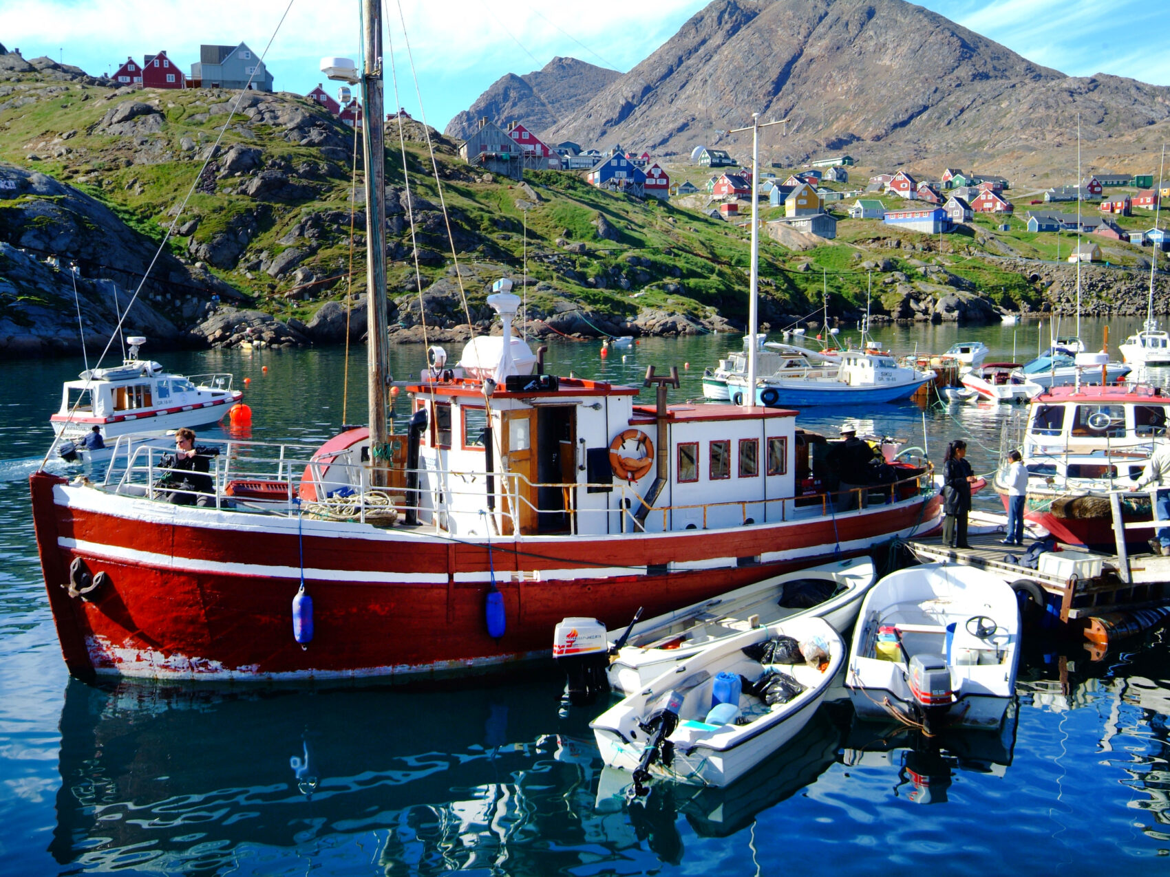 Ostgrönland Hafen mit Fischerbooten in unterschiedlichen Größen, im Hintergrund Berge und grüne Wiesen.