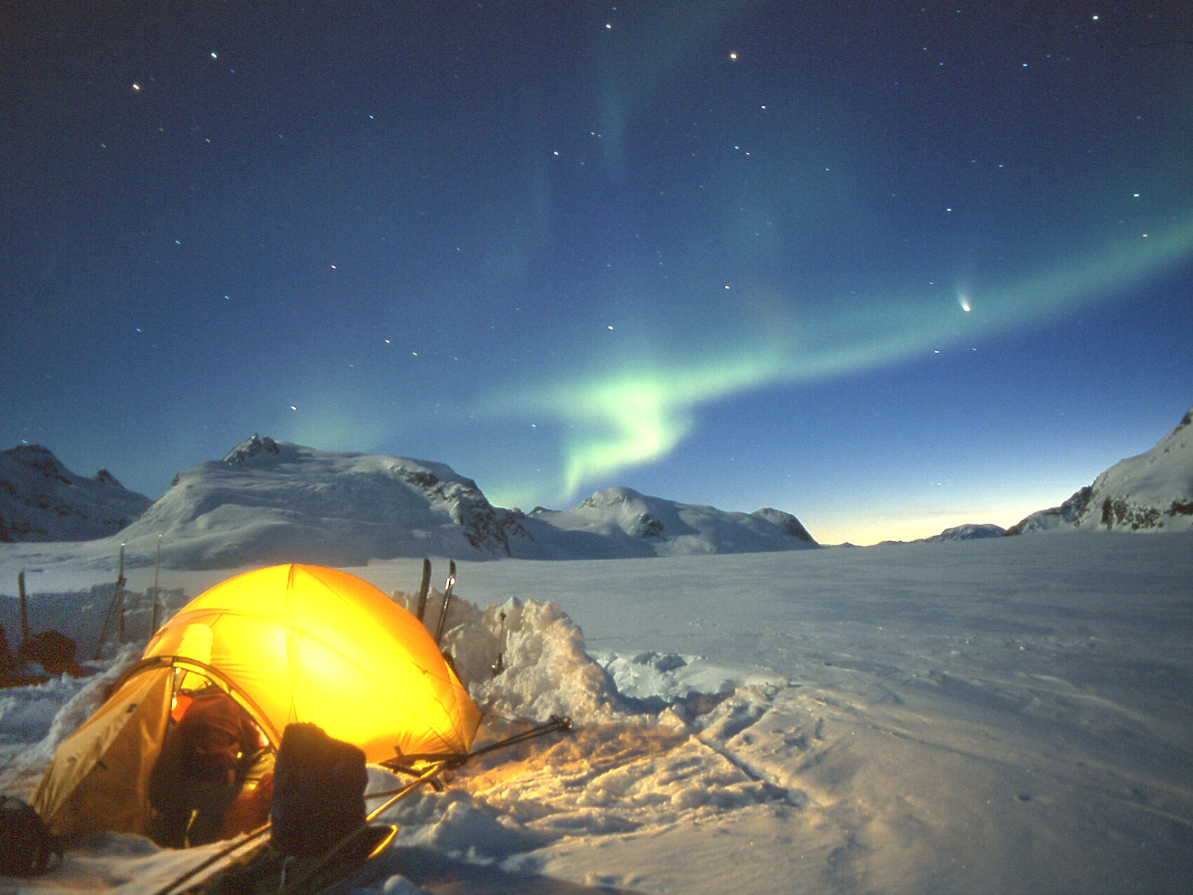 Ostgrönland Nordlichter beim Zeltwandern leuchten in grüner Farbe am Horizont. Zelt leuchtet gelb umgeben von viel Schnee. Sternenklare Nacht. Kleine Berge im Hintergrund.
