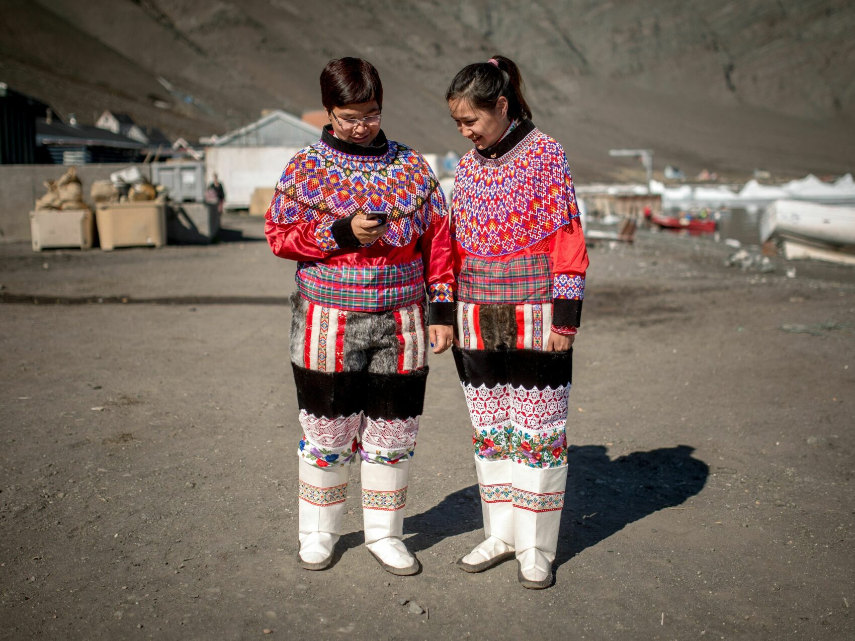 Ostgrönland Illorsuit; zwei Mädchen in nationaler Tracht (sehr bunt mit vielen Mustern) schauen auf ein Handy, im Hintergrund die Ortschaft