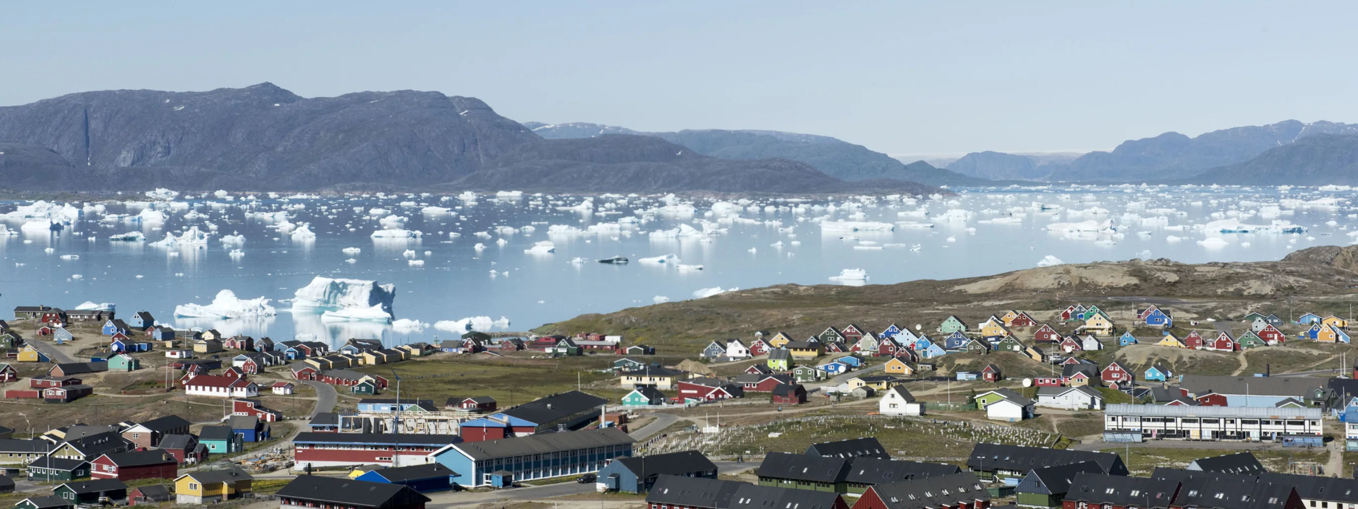 Siedlung mit vielen Häusern in verschiedenen Farben und Fjord mit vielen kleinen Eisbergen im Hintergrund