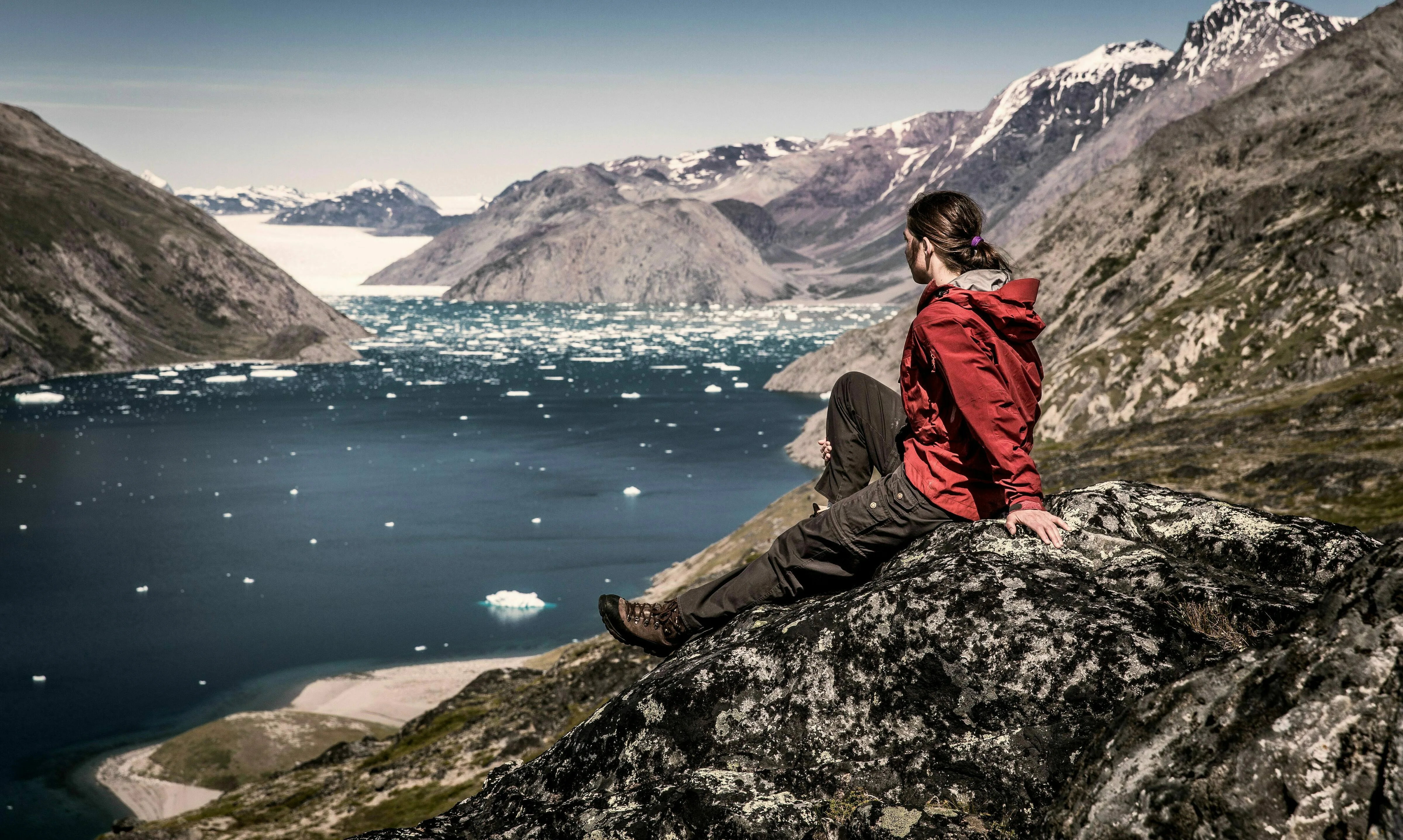 Eine Person sitzt oben auf einem Berg und schaut runter auf einen Eisfjord mit vielen kleinen Eisblöcken