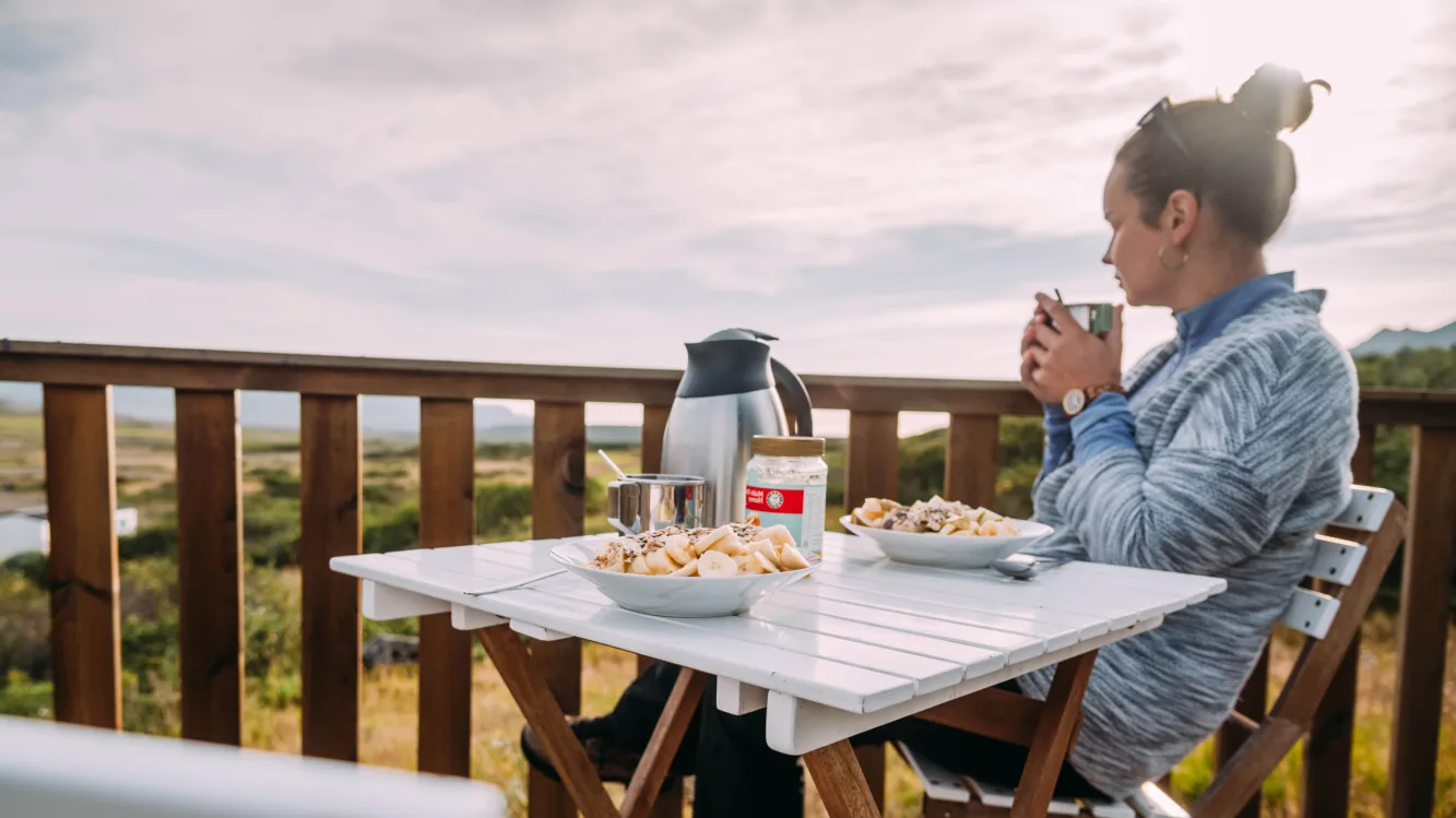 Frau sitzt mit Kaffe in der Hand am Frühstückstisch auf der Terrasse und genießt den Blick auf die weite isländische Landschaft