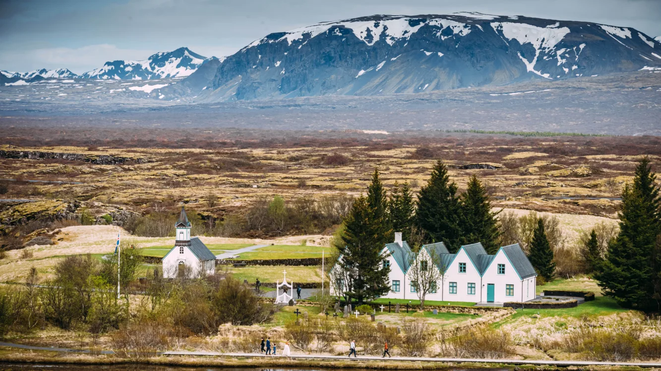 Thingvellir Kirche Island; 
Kirche und 5 weiße Häuser mit grünem Dach, die aneinandergereiht sind
Im Hintergrund kahle und karge Ebene, dahinter Berge leicht mit Schnee bedeckt