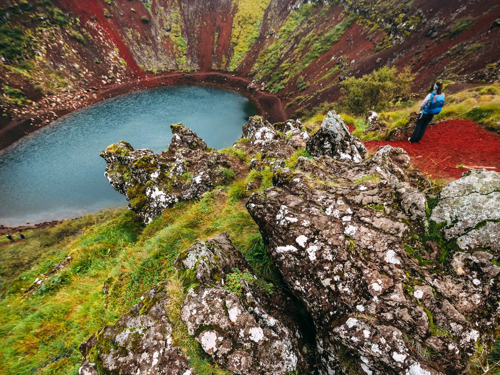 Person steht oben an einem Krater mit Blick auf den Kratersee, mit grünem Islandmoos und rötlichem Gestein drumherum