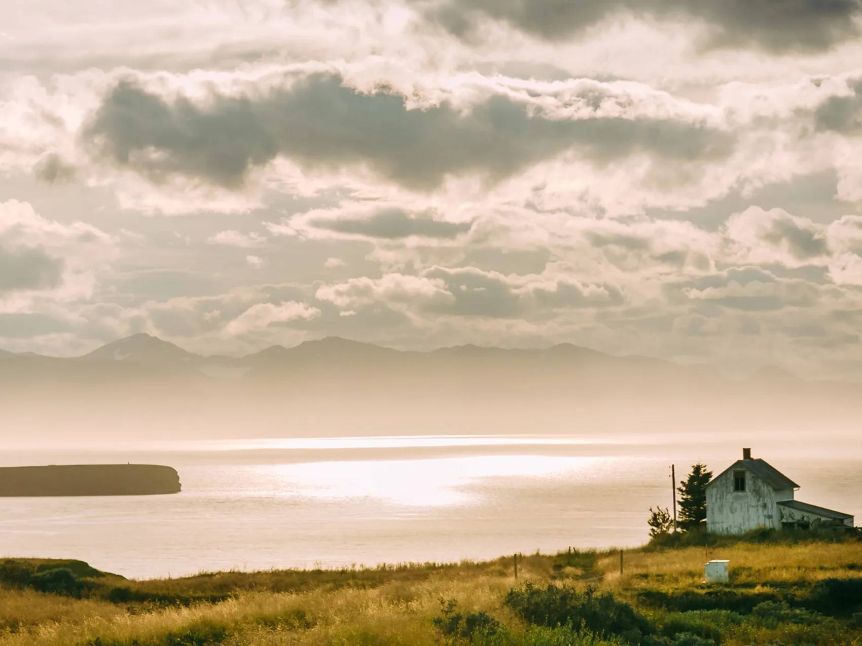 Ein kleines Haus direkt am Meer bei Sonnenuntergang, im Hintergrund sind Berge und Wolken zu sehen