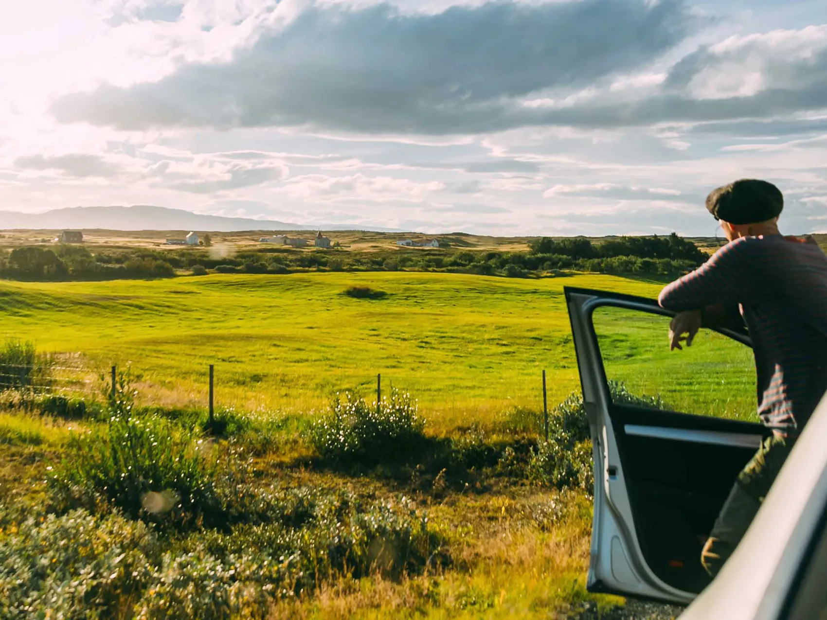 Mann lehnt sich an der Autotür an und beobachtet die schöne Aussicht auf eine große grüne Wiese mit mehreren kleinen Häusern im Hintergrund