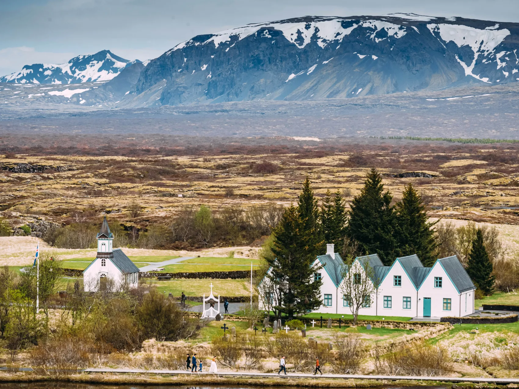 Thingvellir Kirche Island; 
Kirche und 5 weiße Häuser mit grünem Dach, die aneinandergereiht sind
Im Hintergrund kahle und karge Ebene, dahinter Berge leicht mit Schnee bedeckt