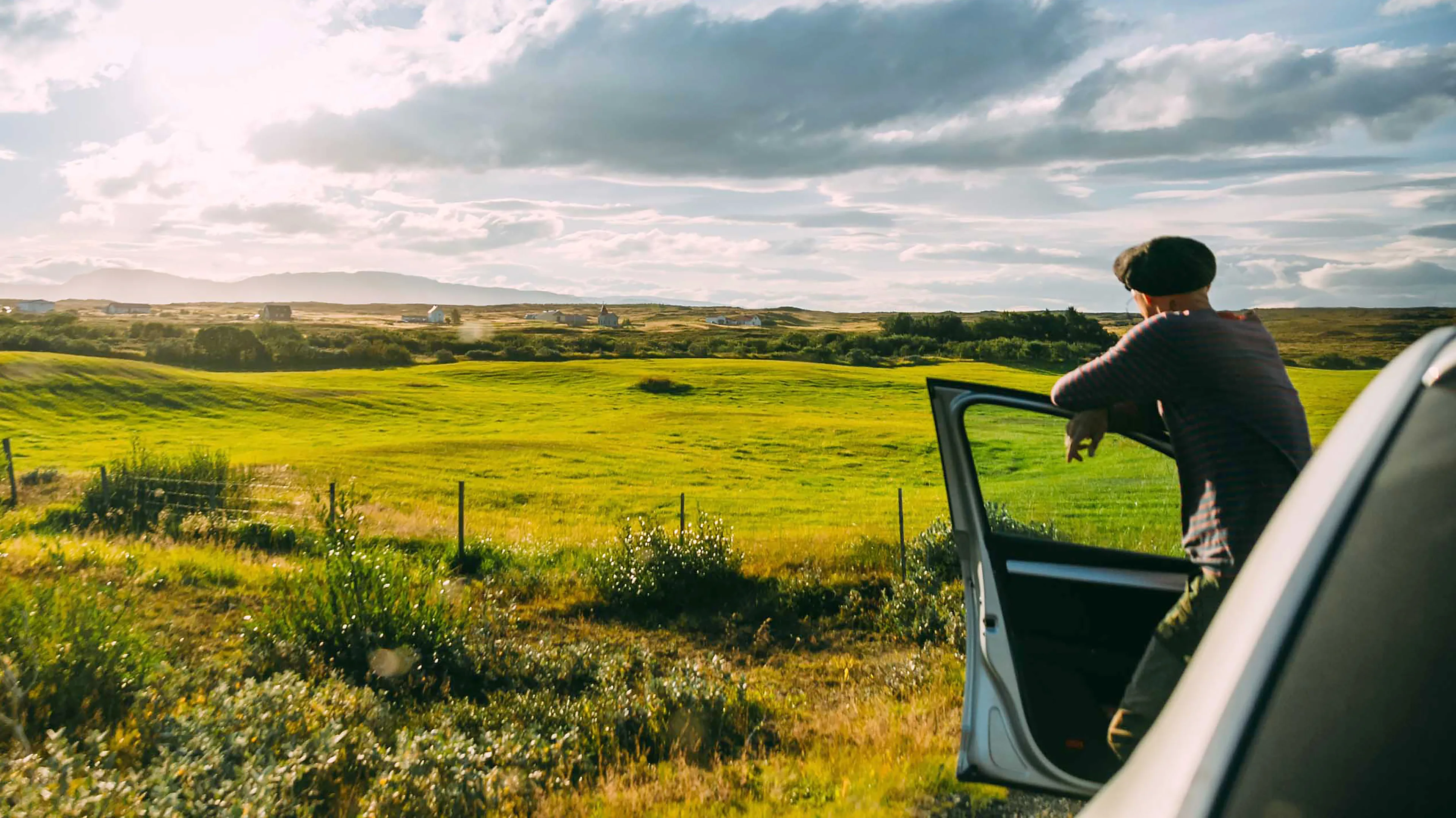 Mann lehnt sich an der Autotür an und beobachtet die schöne Aussicht auf eine große grüne Wiese mit mehreren kleinen Häusern im Hintergrund