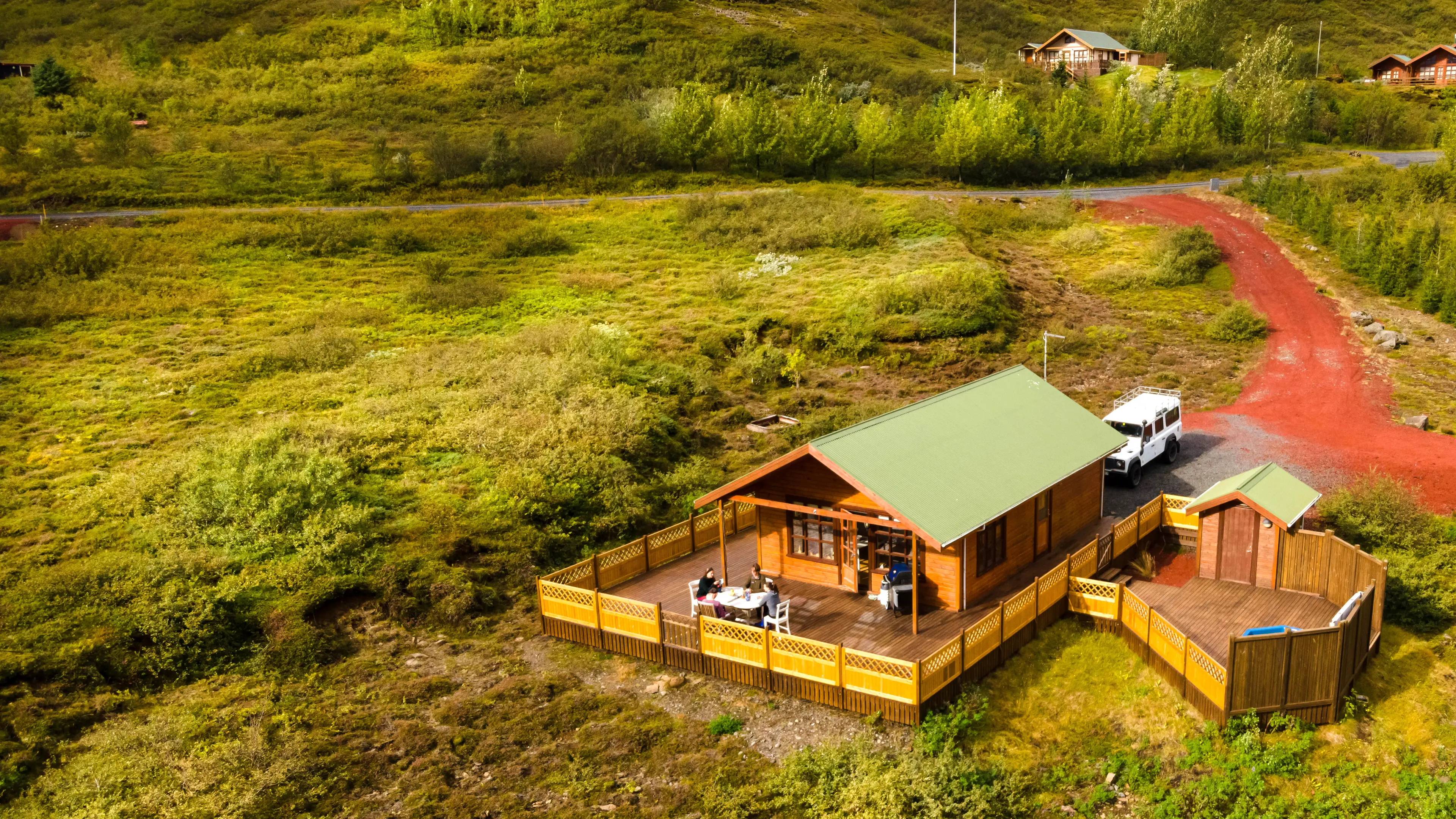 Luftaufnahme eines Ferienhauses, vier Leute sitzen auf der Terasse, ein weißer Jeep steht vor dem Haus, umherum weite grüne Landschaft und ein roter Weg der zum Haus führt