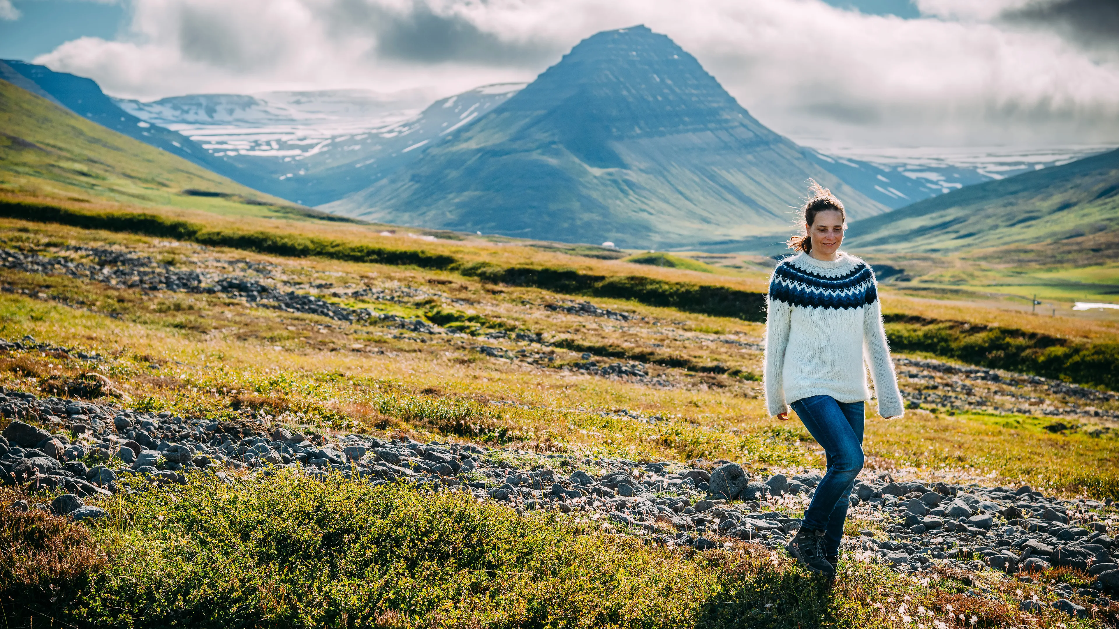 Eine Frau in Islandpullover wandert einen Berg hoch, im Hintergrund sind weitere berge und Wolken zu sehen