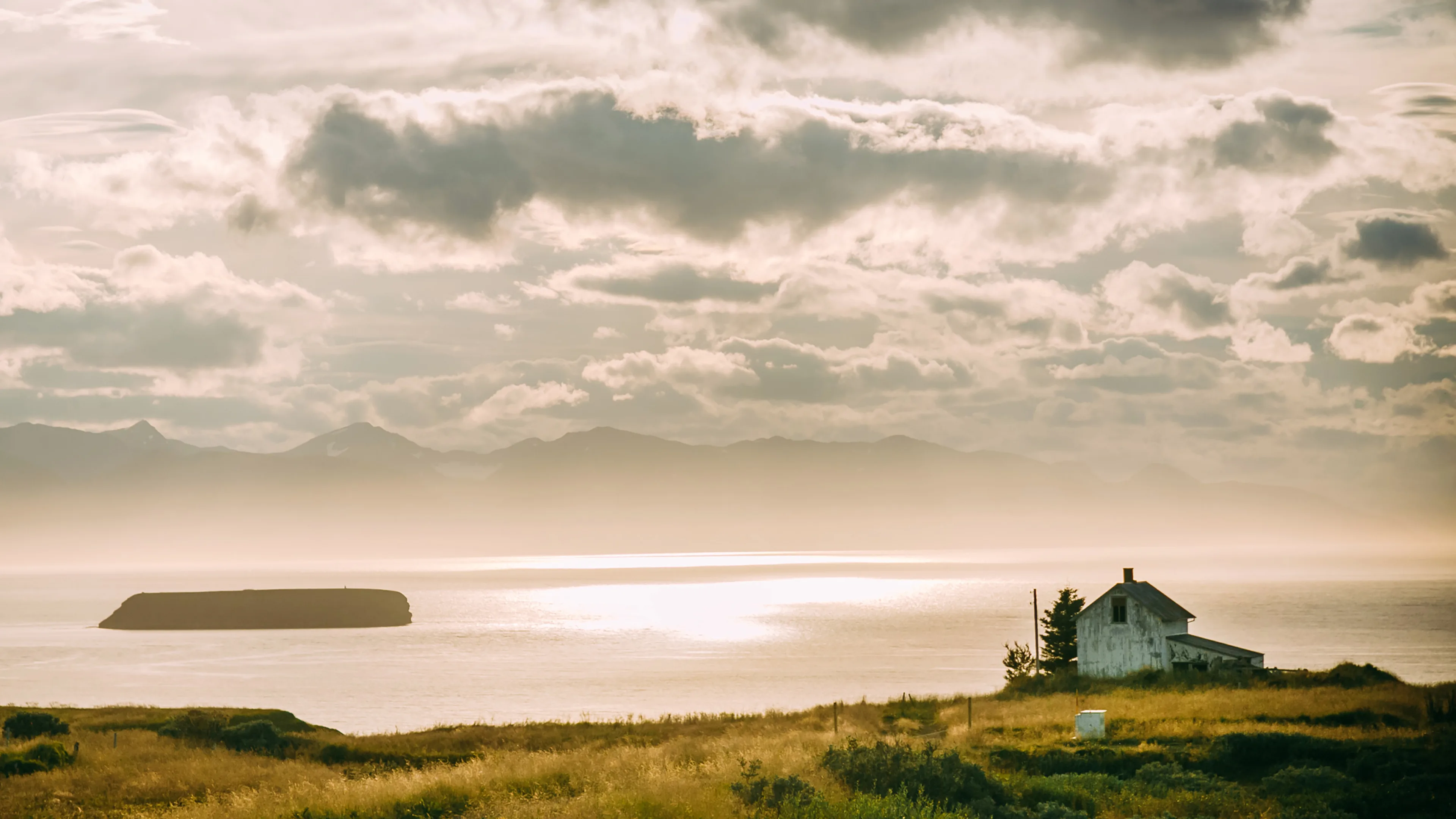 Ein kleines Haus direkt am Meer bei Sonnenuntergang, im Hintergrund sind Berge und Wolken zu sehen