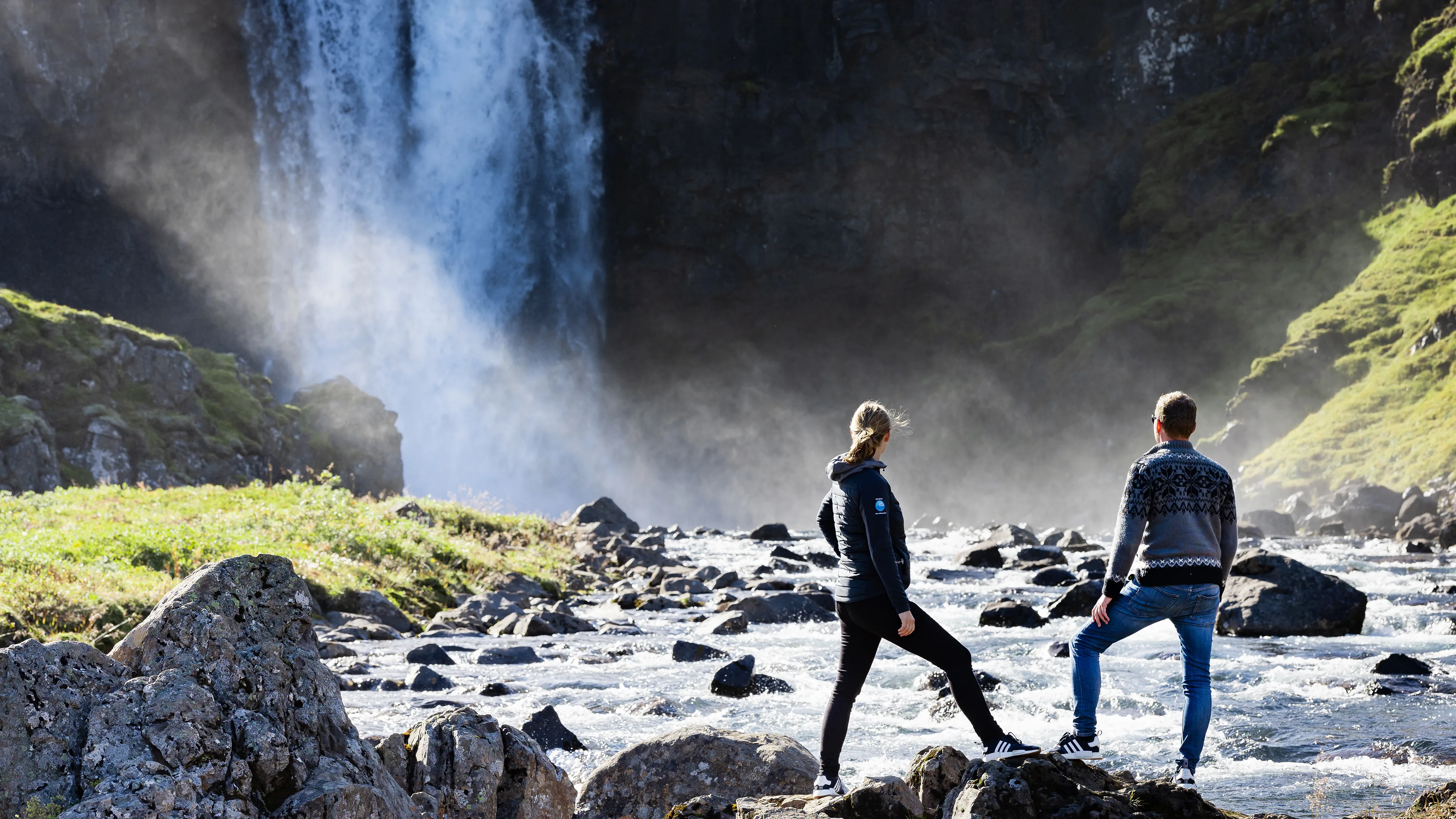 Paar am Wasserfall auf Island, Sprüh-Nebel durch herabstürzendes Wasser, Sonnenstrahlen, kleine Felsen im Wasser