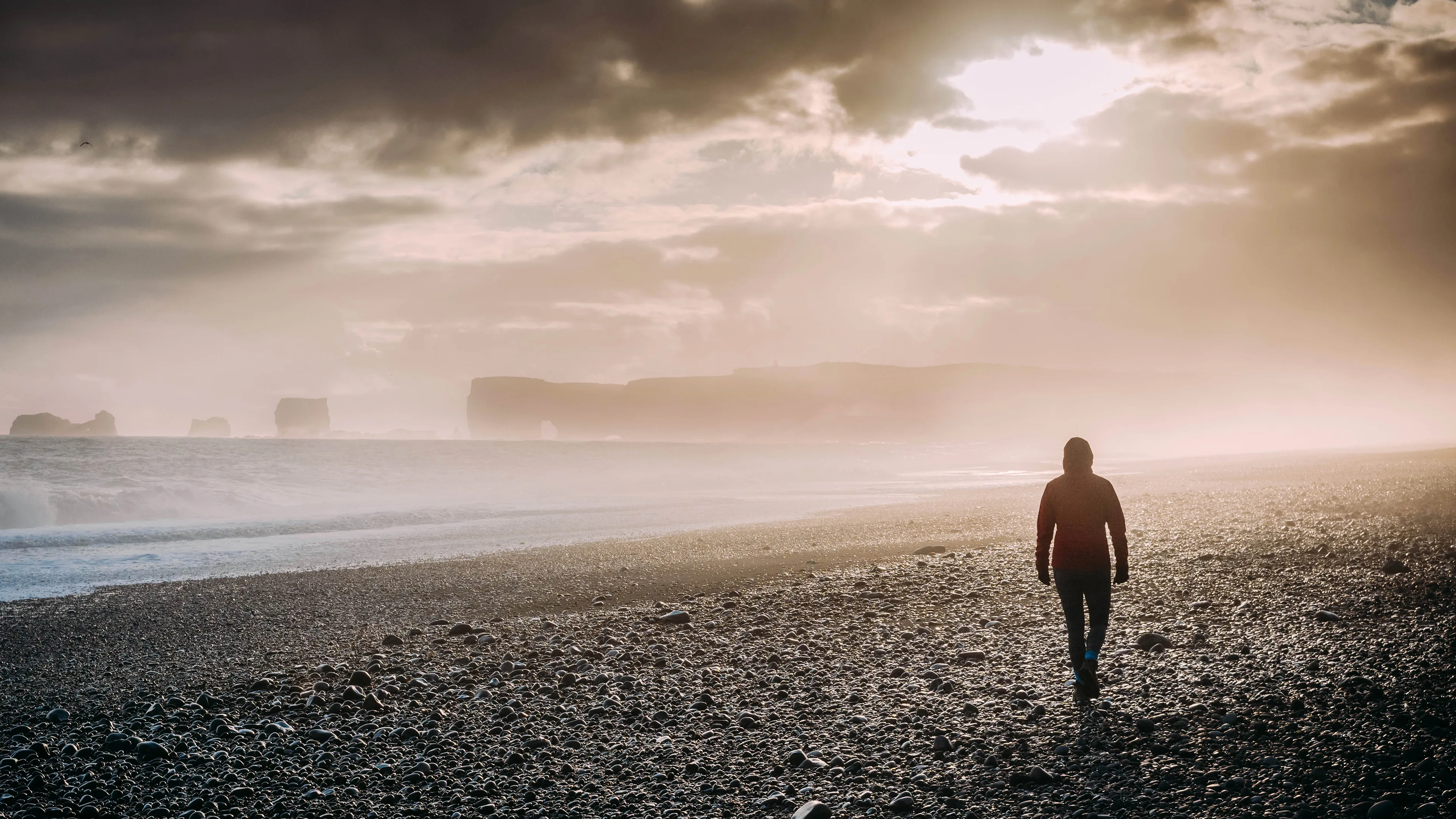 Person am Reynisfjara Strand auf Island, Nebel, Sonne bricht durch dunkle Wolkendecke, Felsenküste mit einzigartigen Formationen im Hintergrund