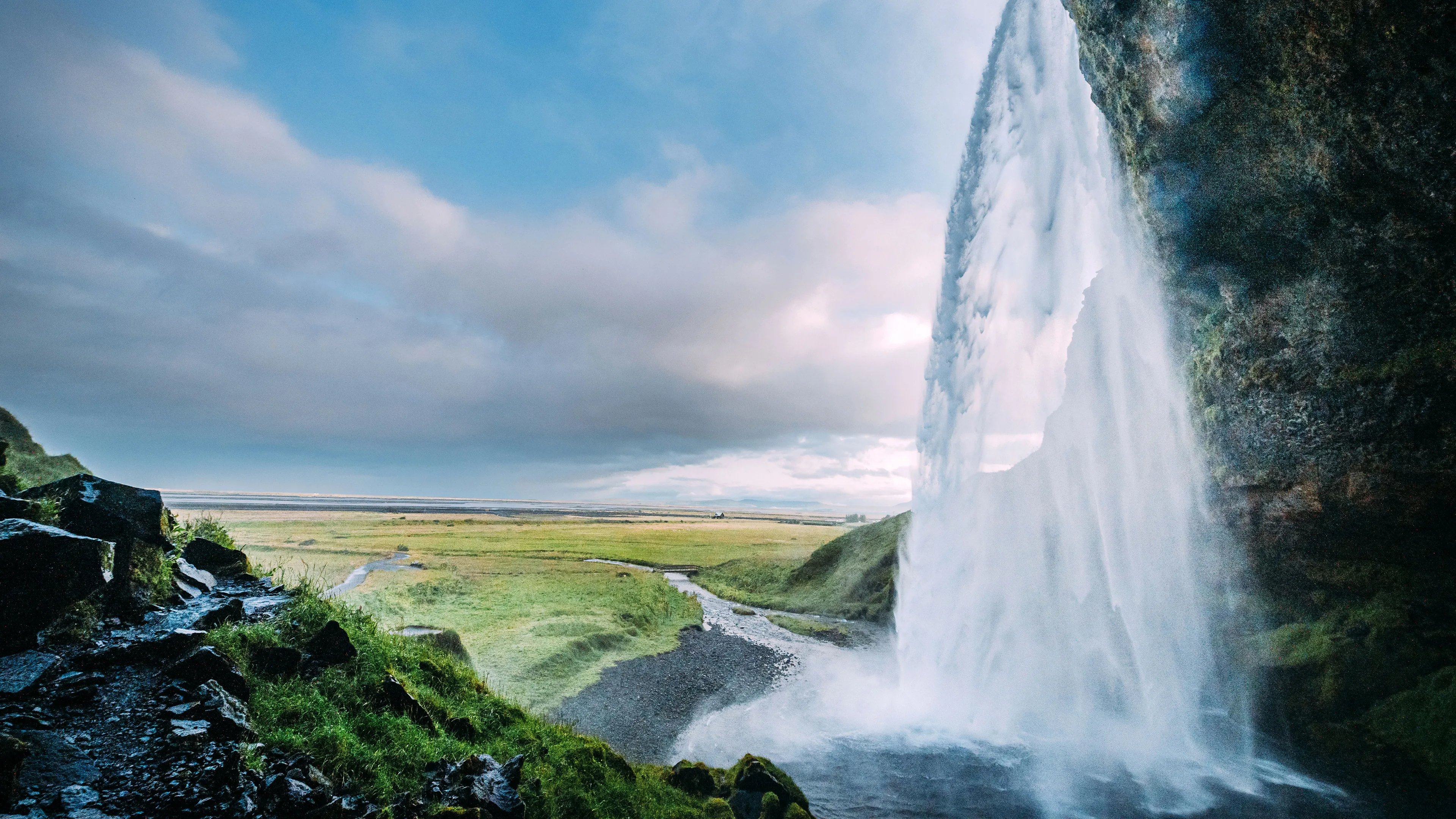 Seljalandsfoss Wasserfall Island, Blick von hinter dem Wasserfall auf Landschaft davor, blauer Himmel mit Wolken, grüne weite Graslandschaft