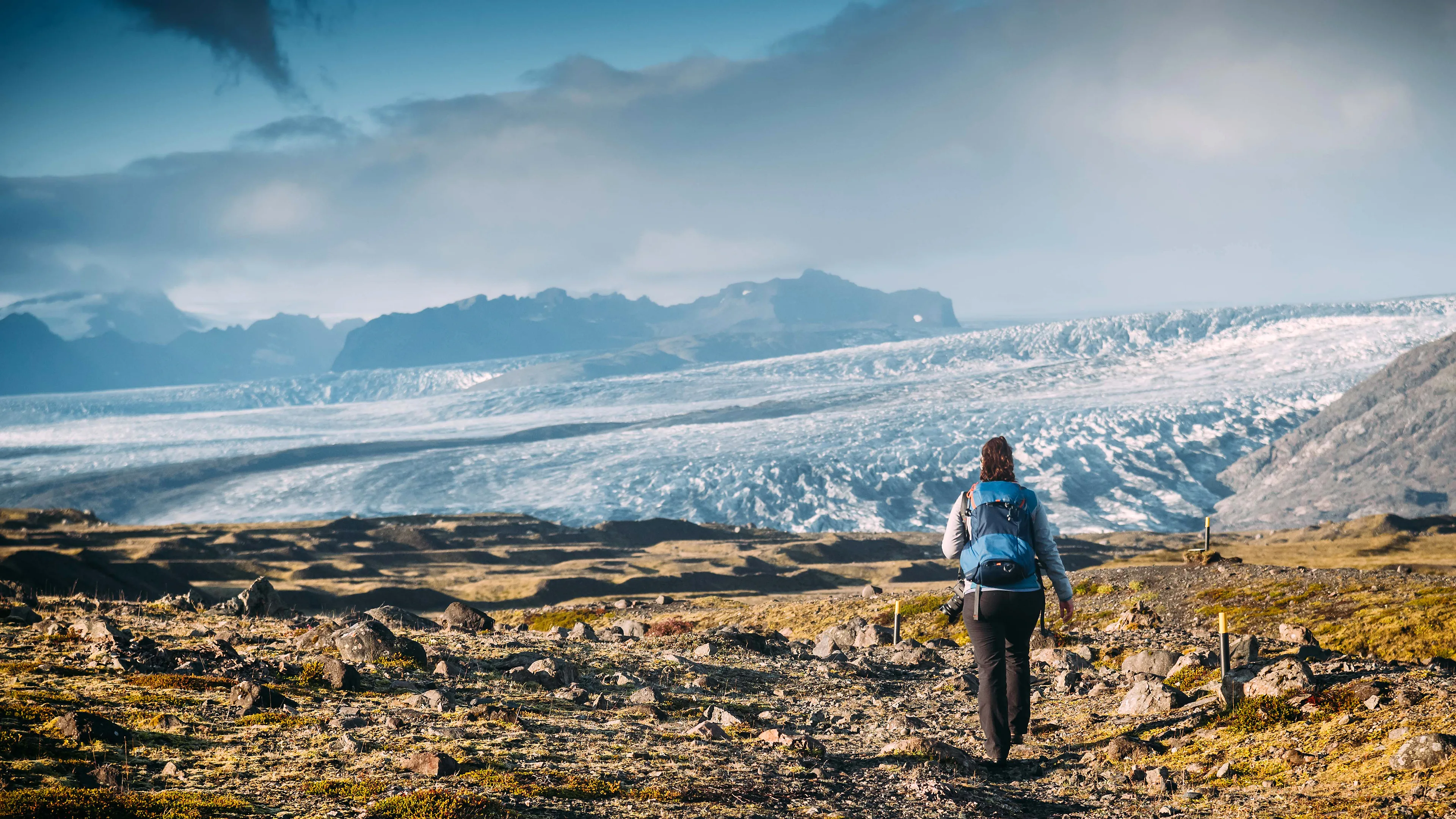 Eine Frau wandert auf eine große Gletscherzunge zu, im Hintergrund Berge und bewölktes Wetter