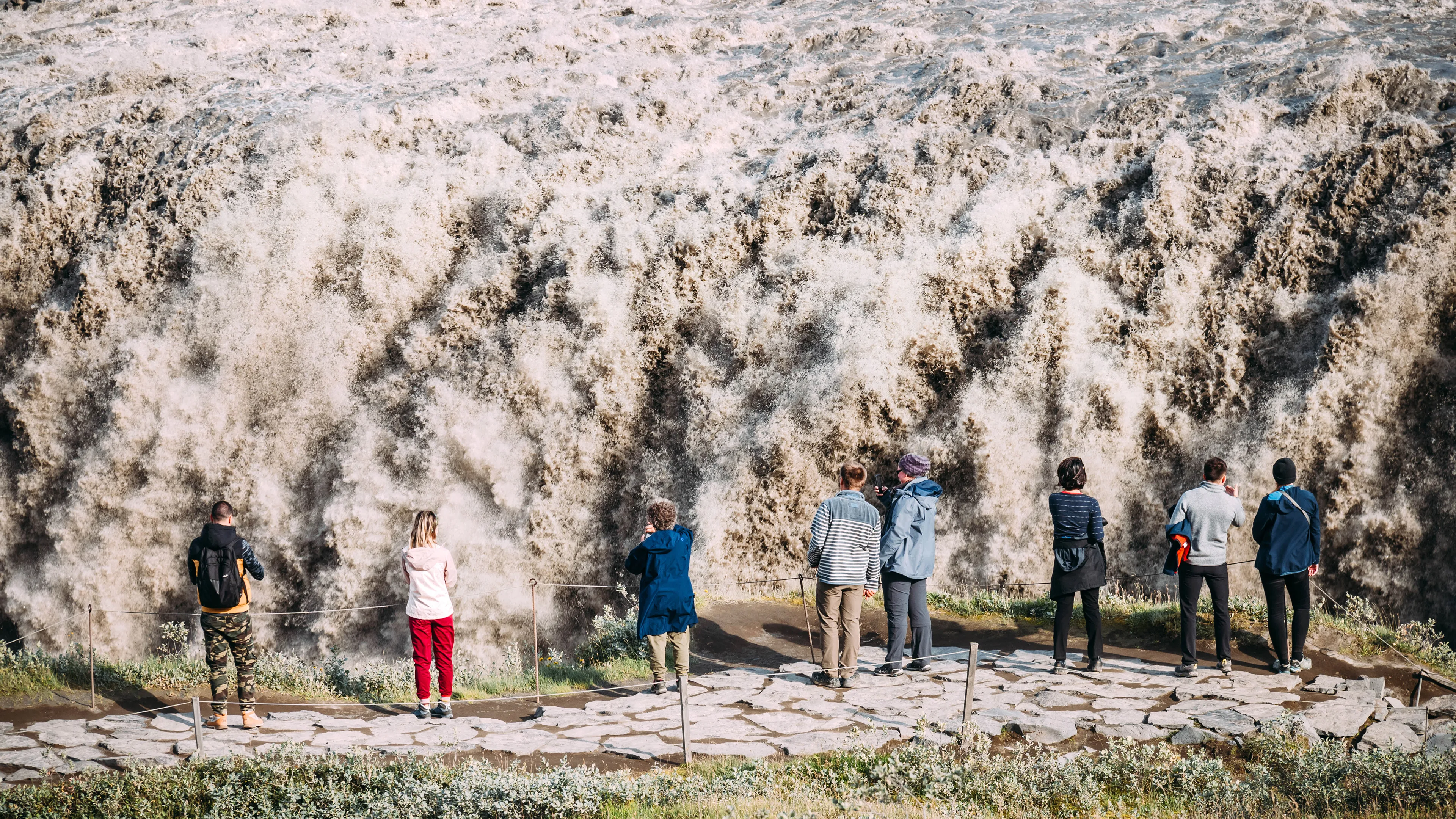 Mehrere Touristen stehen vor dem großen Wasserfall Dettifoss und fotografieren diesen