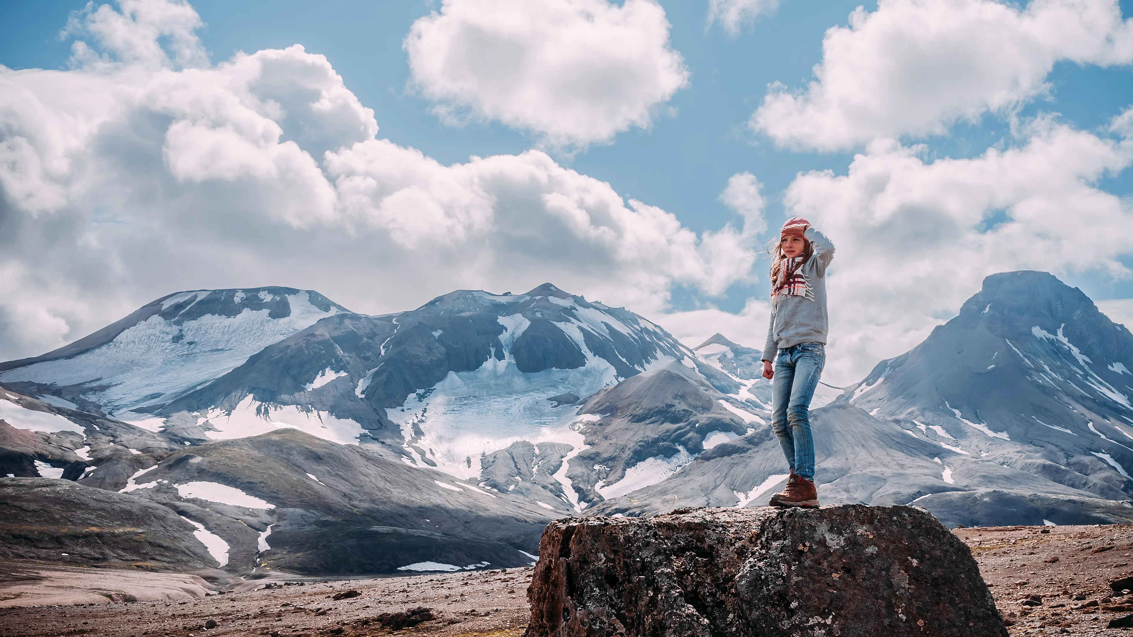 Mädchen steht auf einem kleinen Felsen und beobachtet die Landschaft, im Hintergrund große verschneite Berge und Wolken