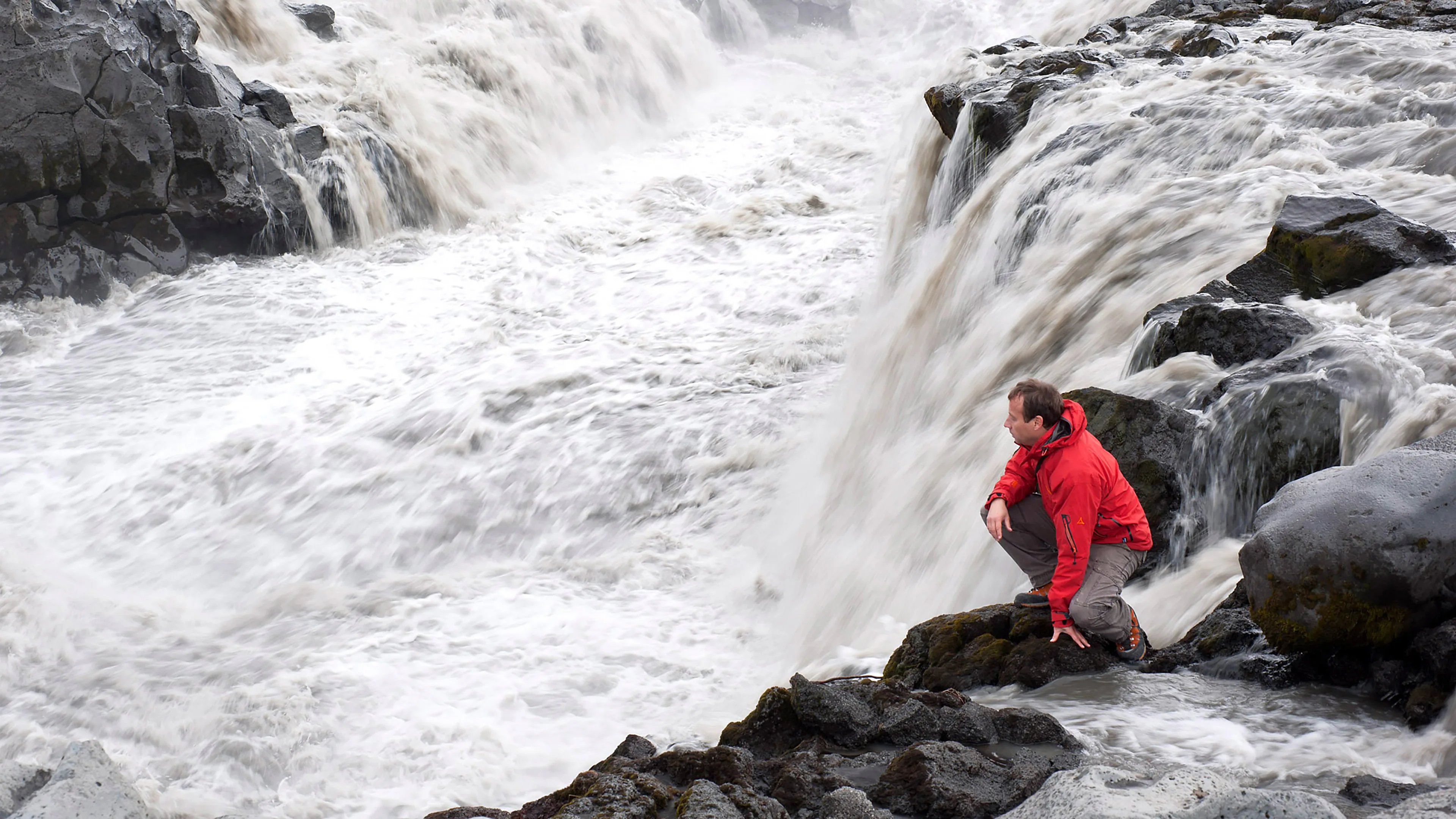 Mann in roter Jacke kniet direkt neben einem Wasserfall im isländischen Hochland