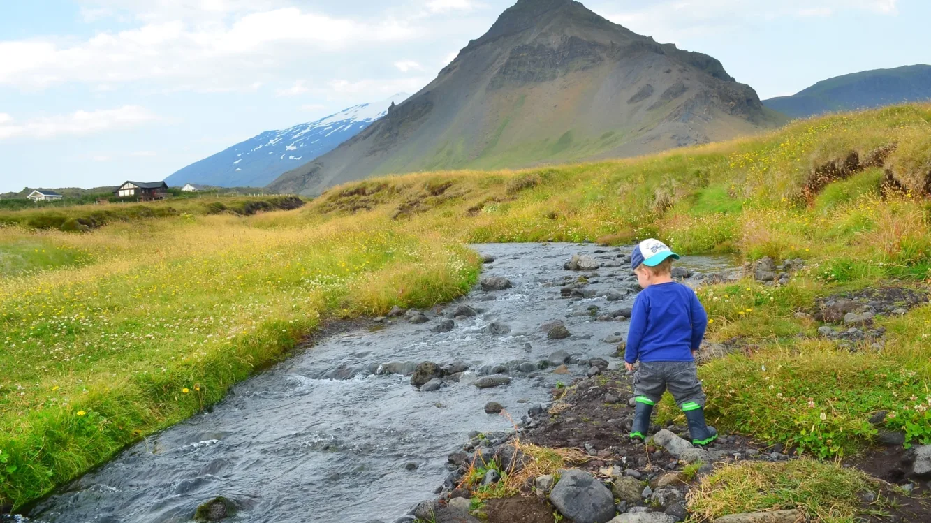 Kind spielt am Bach in Island, Berge im Hintergrund, saftig grüne Wiese, blauer Himmel mit Wolken