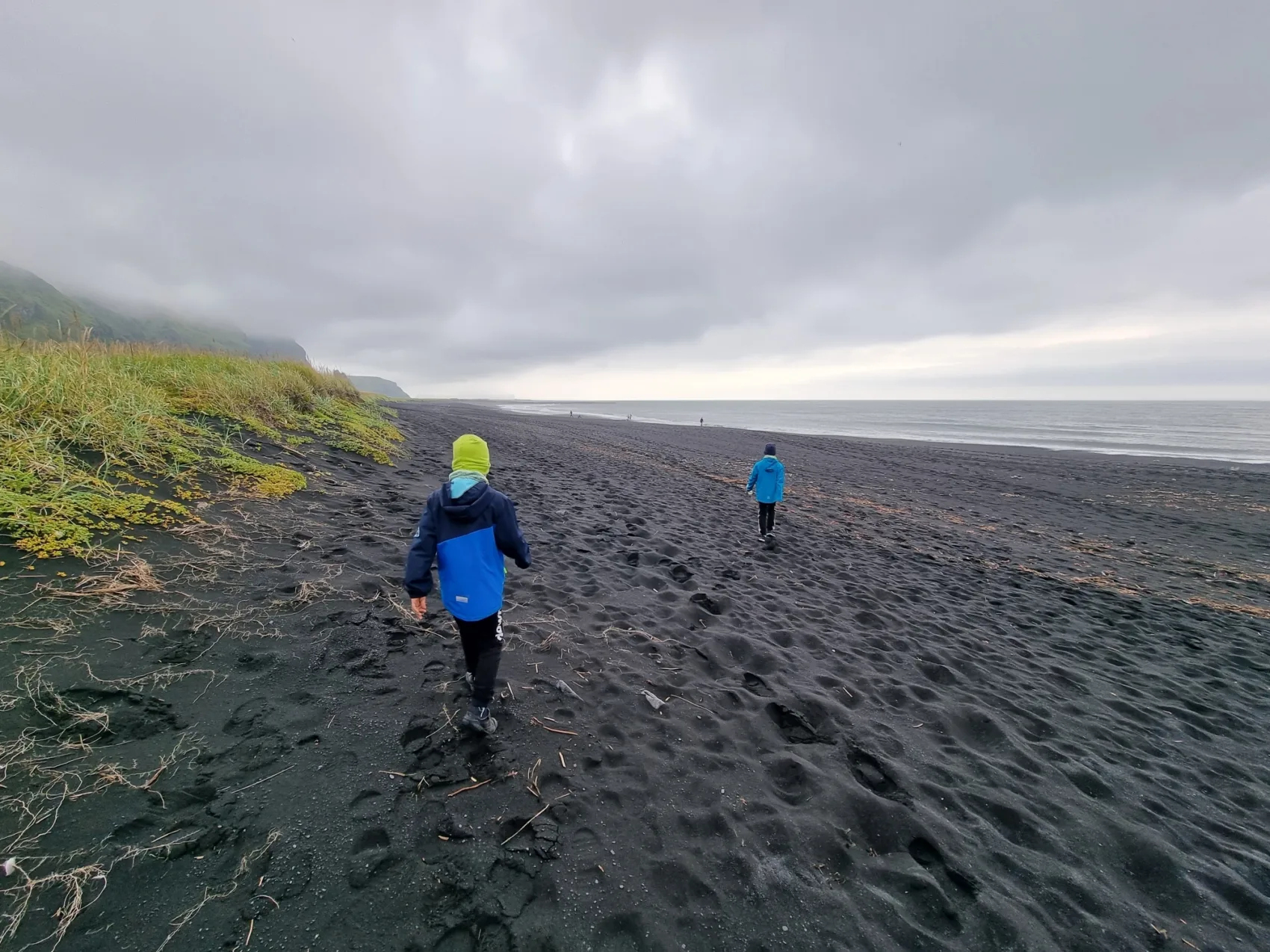 Zwei Kinder am Strand von Vik, schwarzer Sandstrand, grauer Wolkenhimmel, Spuren im Sand, ruhiges Meer