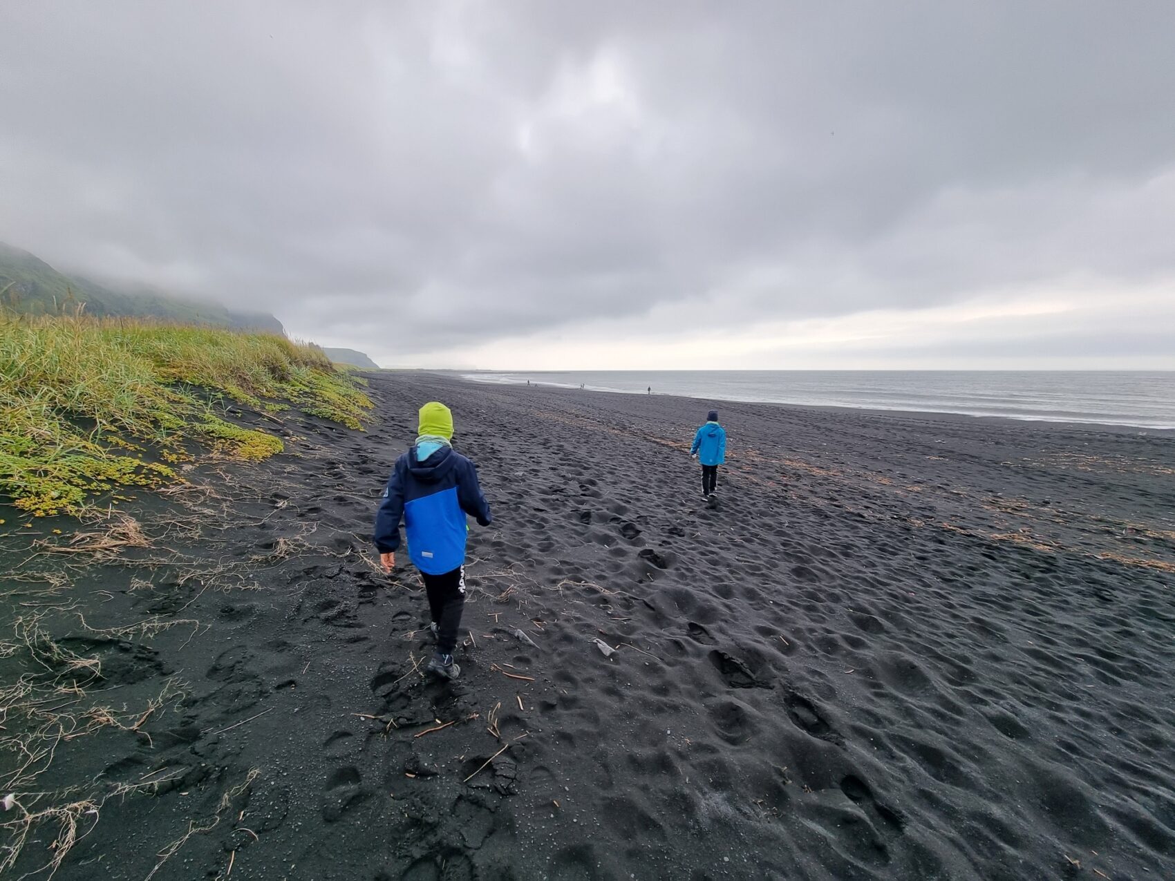 Zwei Kinder am Strand von Vik, schwarzer Sandstrand, grauer Wolkenhimmel, Spuren im Sand, ruhiges Meer