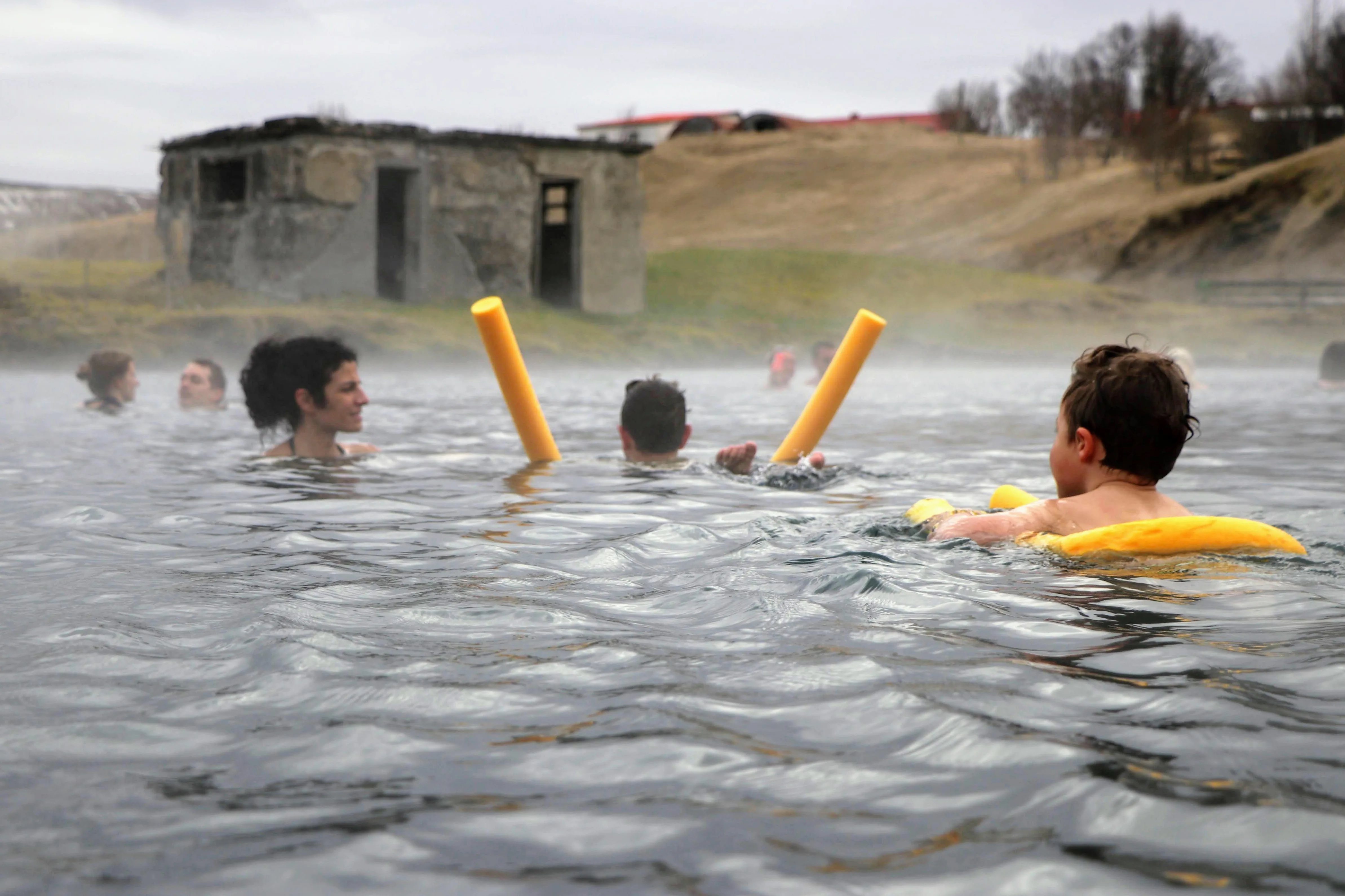 Kinder baden in der Secret Lagoon in Island, im Hintergrund eine alte Steinhütte