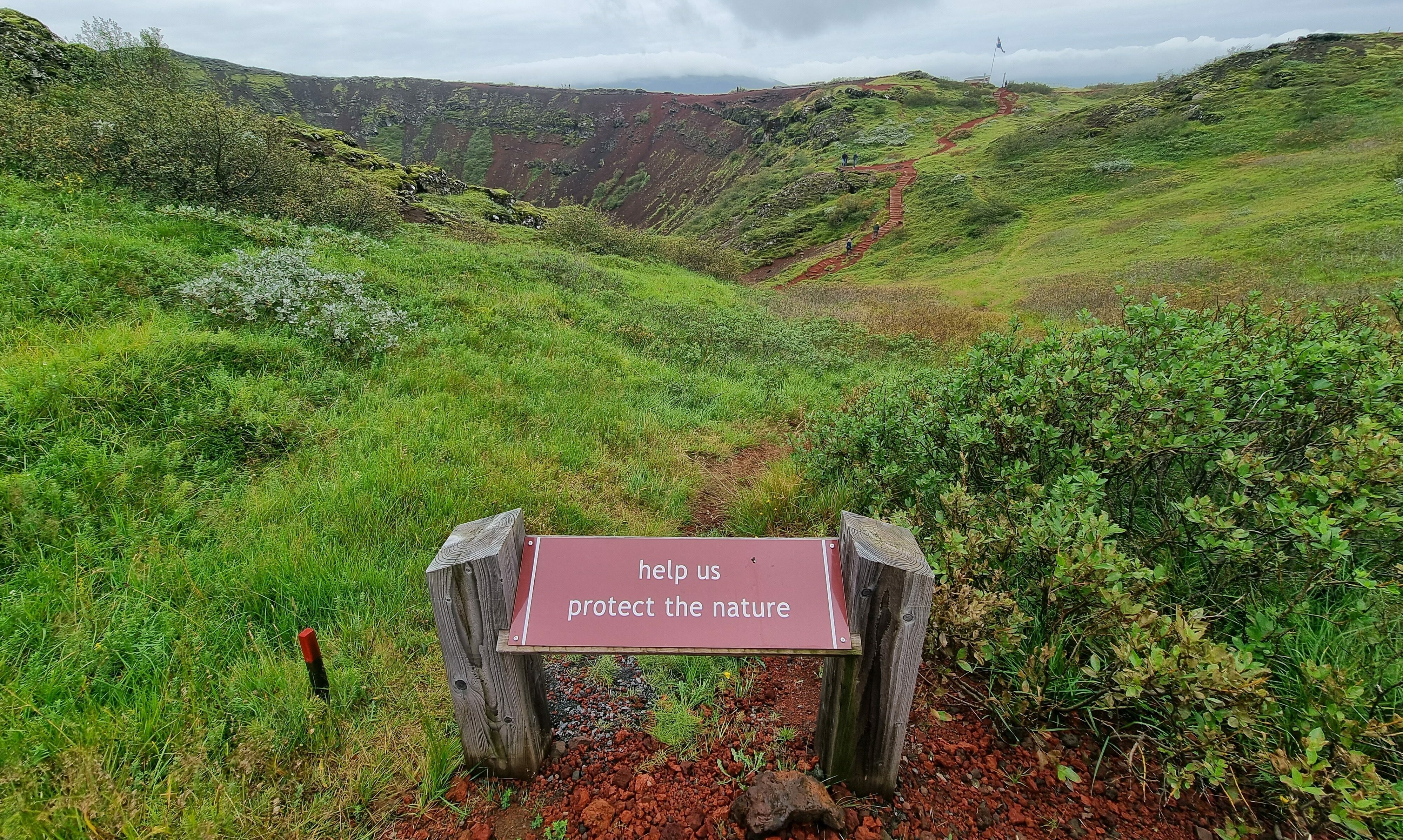 Naturschutz - help us protect nature - Schild in rot vor einem mit Gras bewachsenen Krater/Wandergebiet