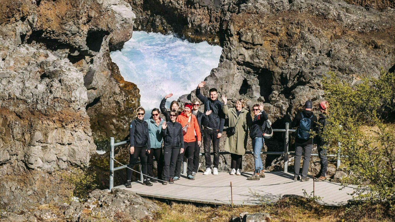 Katla Team am Hraunfossar, das Team steht auf einer Plattform vor dem Wasserfall und hebt die Hände - sie sind von Lavafelsen umgeben