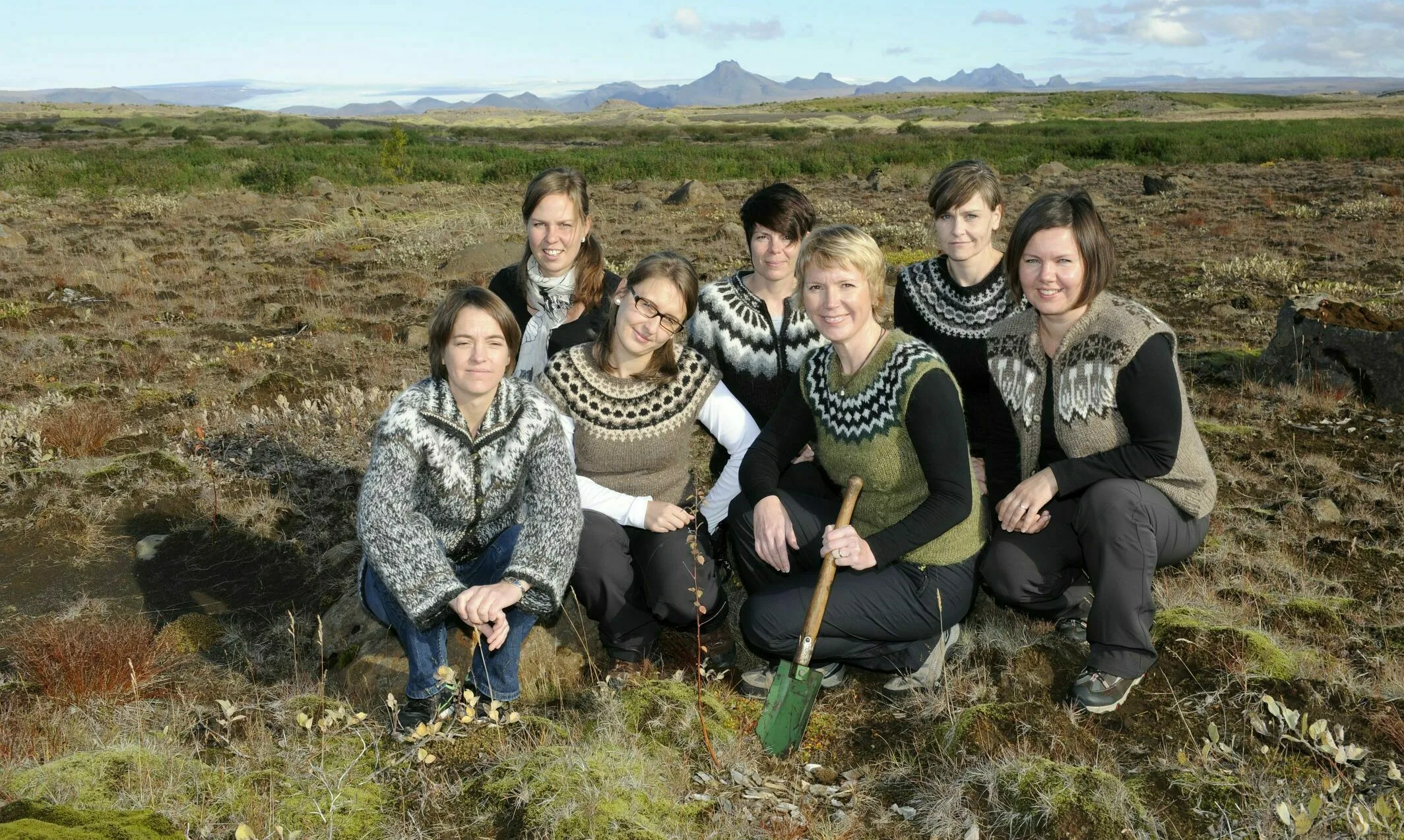 Katla Team in Island - Bäume pflanzen
Sieben Mitarbeiterinnen des Katla Teams Knien am Boden, eine hält eine Schaufel in der Hand. Im Hintergrund die Hochebene zu erkennen.