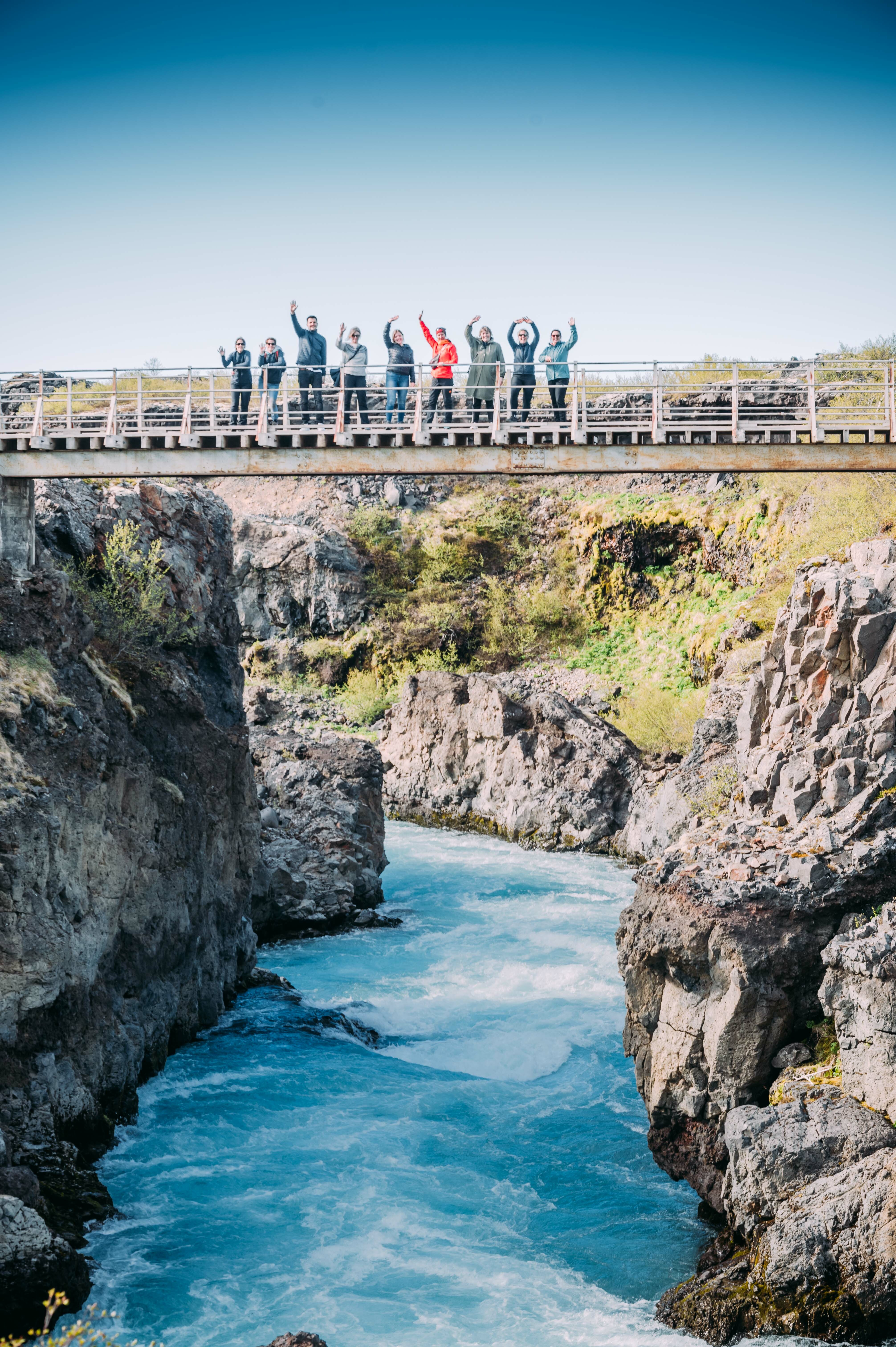 Katla Team beim Hraunfossar, stehen in der Mitte der Brücke und heben die Hände, unter ihnen ein türkisblauer Fluss.