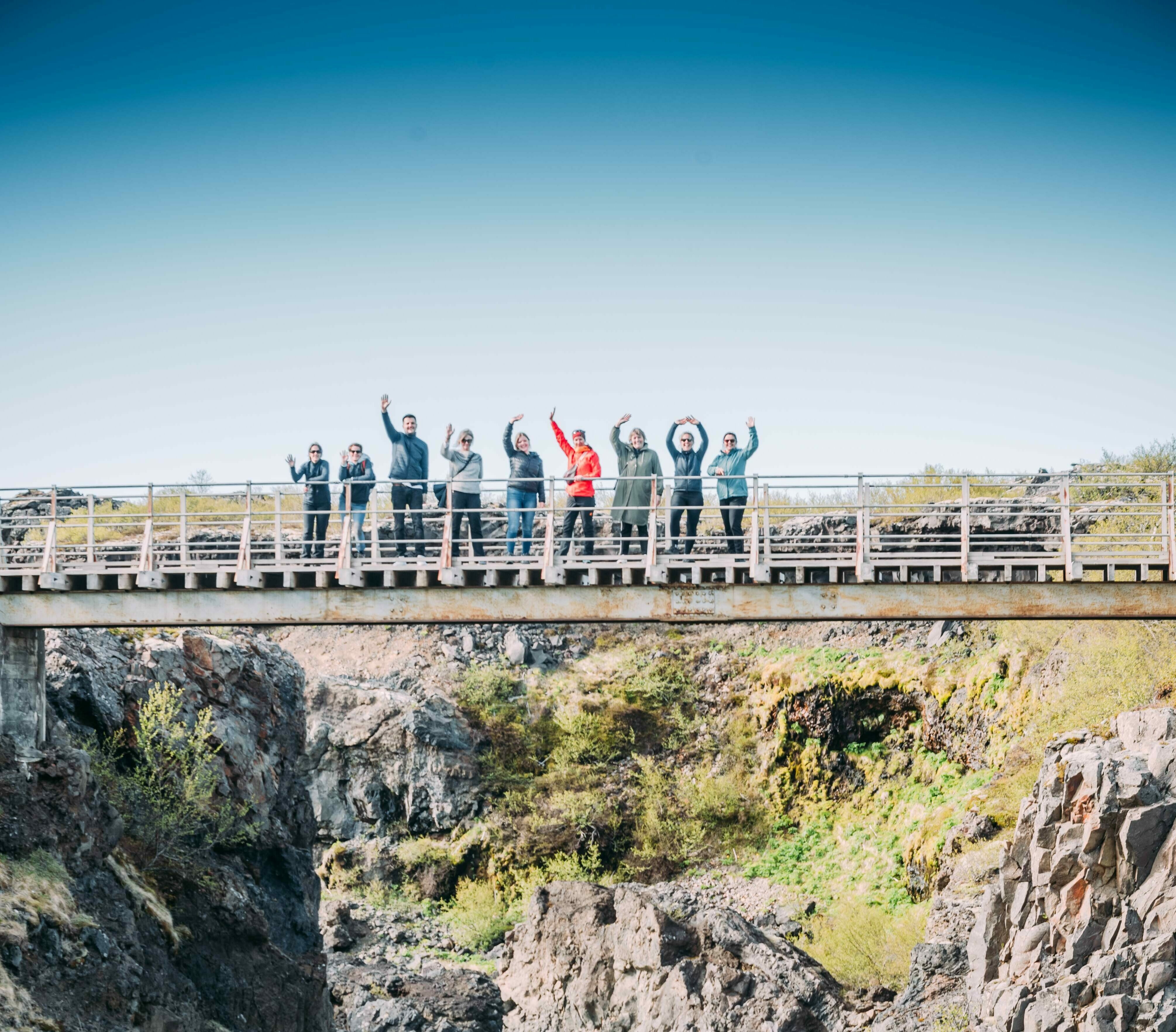 Katla Team winkend nebeneinander auf der Eisenbrücke am Hraunfossar Wasserfall