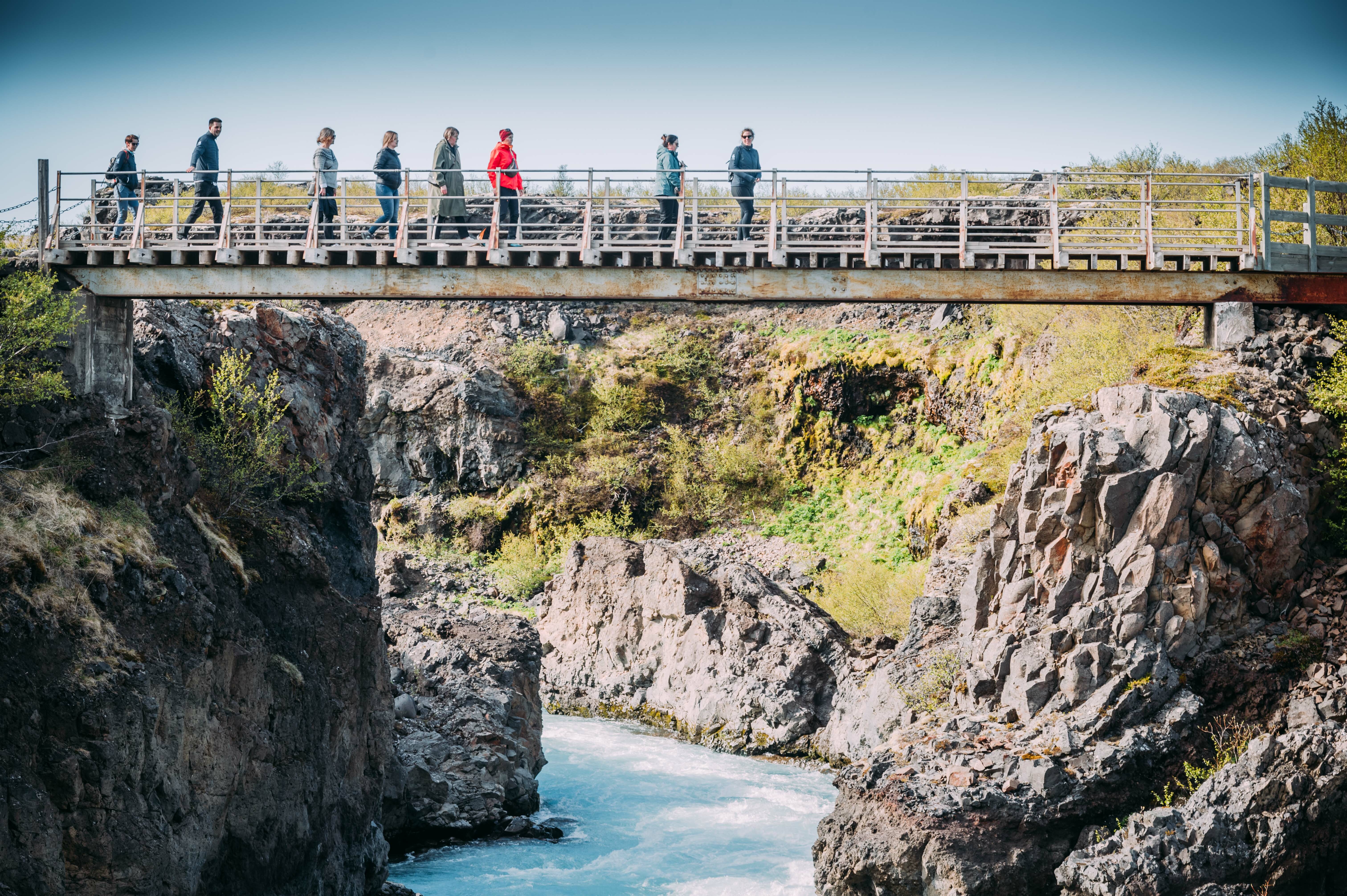 Katla Team Hraunfossar, das Team läuft über eine Brücke, unter ihnen eine Schlucht mit Fluss, Felswände an den Seiten