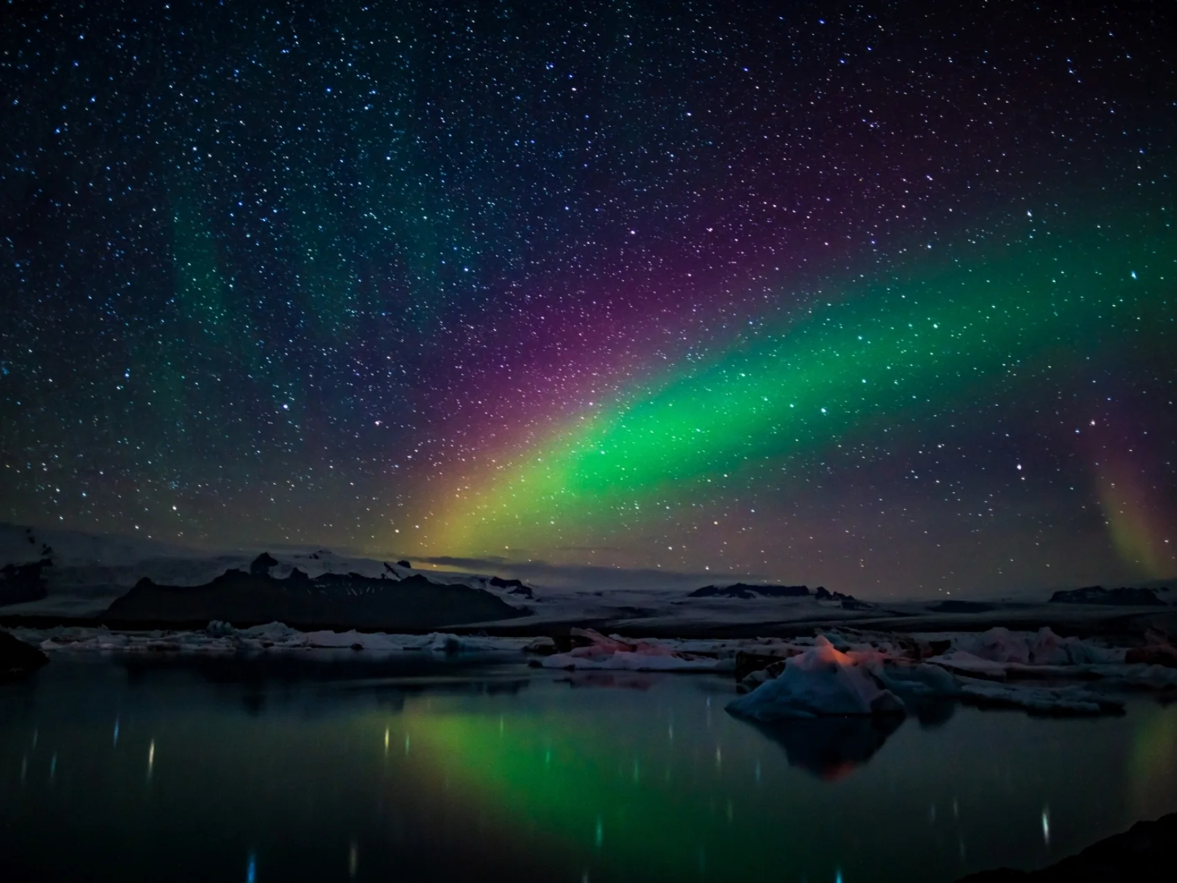 Nordlichter Island 
Über einem Gewässer bei sternenklarer Nacht sieht man die Polarlichter in geld, grün und pink leuchten. Im Hintergrund sind mit Schnee bedeckte Berge.