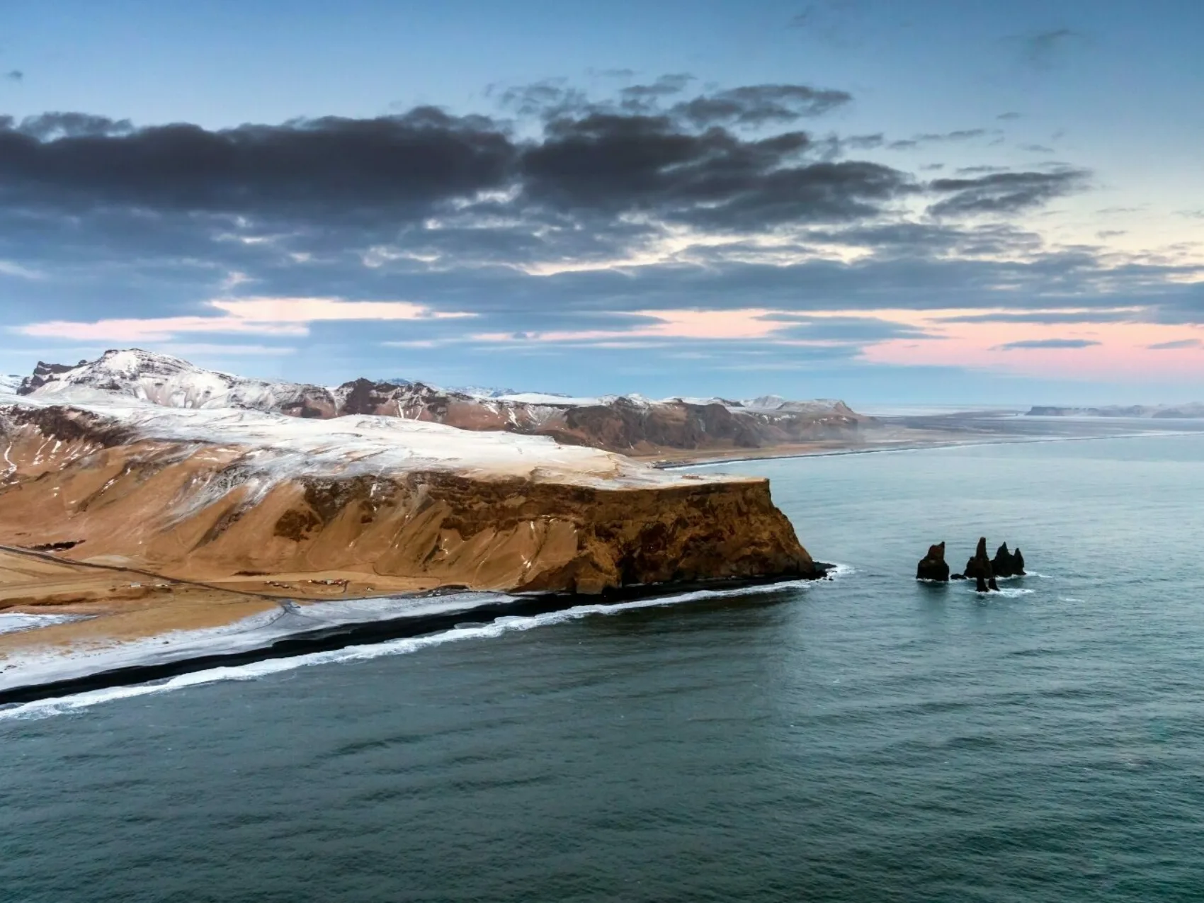 Reynisfjall Küste, Südisland mit Schnee bedeckt, schwarzer Strand und Felsen im Wasser, dunkle Wolken im Hintergrund