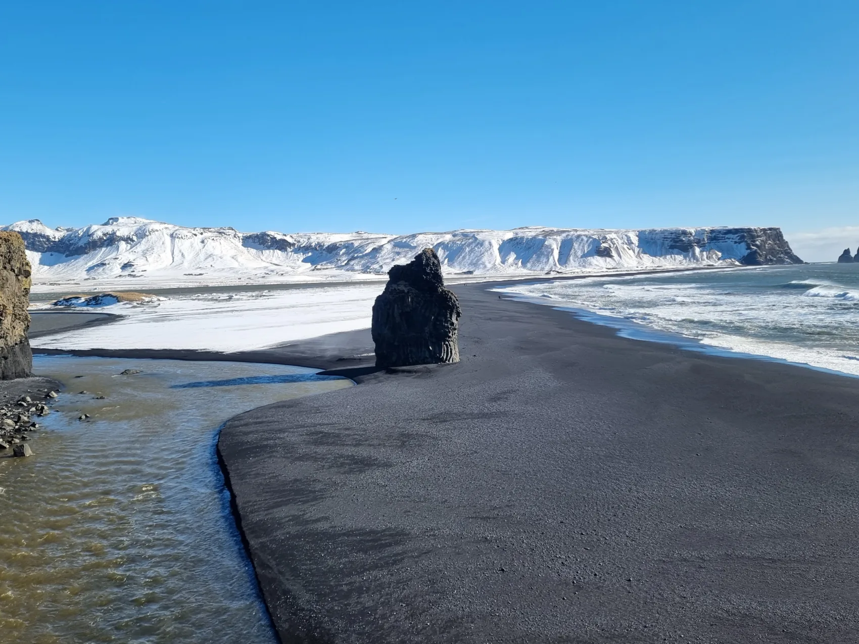 Der Strand von Dyrhólaey im Süden Islands mit schneebedeckten Bergen im Hintergrund.