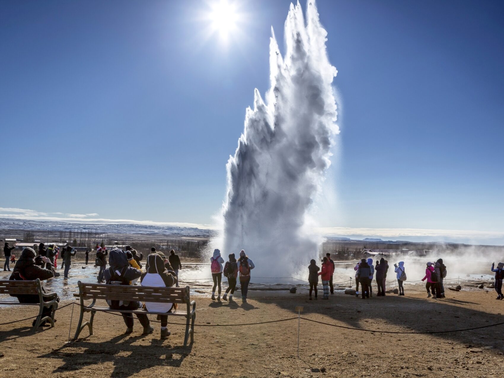 Geysir Strokkur Island, viele Menschen schauen dem ausbrechenden Geysir Strokkur bei Sonnenschein zu. Ein Wasser und Dampfwolke wird in den Himmel geschossen.