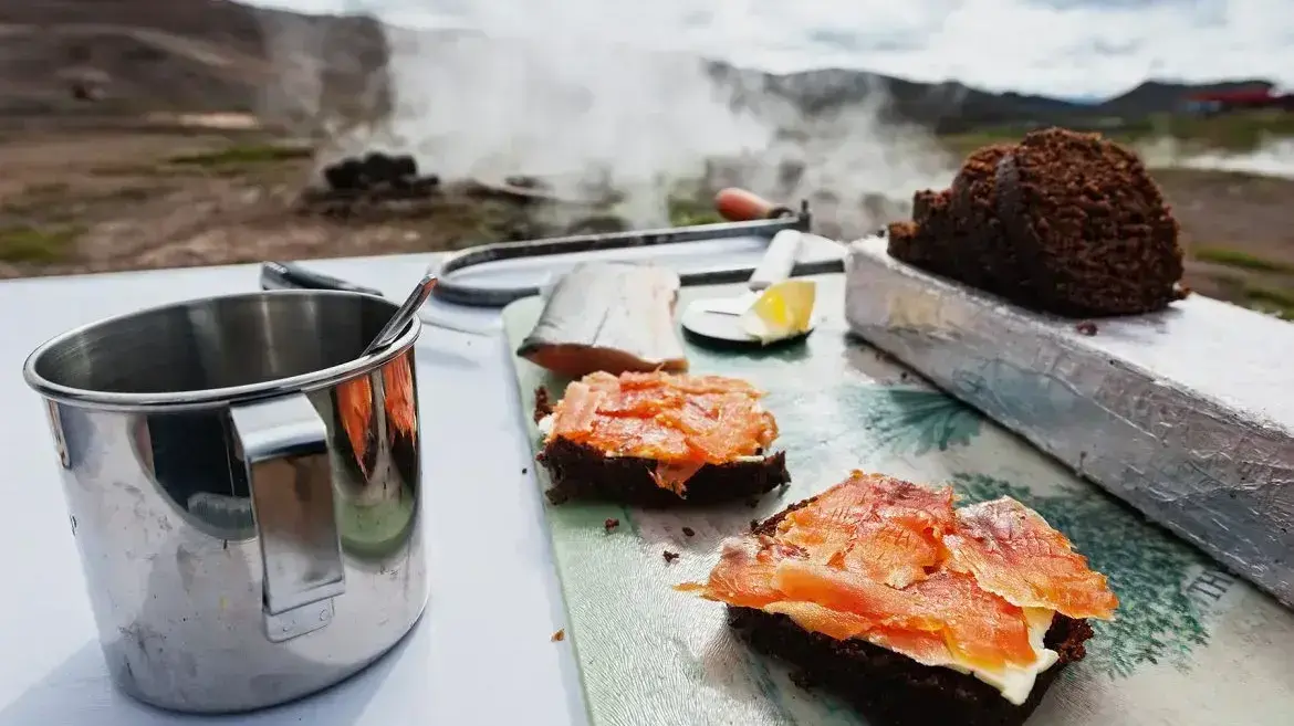Ein Tisch mit einem isländischen Erdbrot mit Lachs obendrauf, und einer heiß dampfenden Quelle im Hintergrund