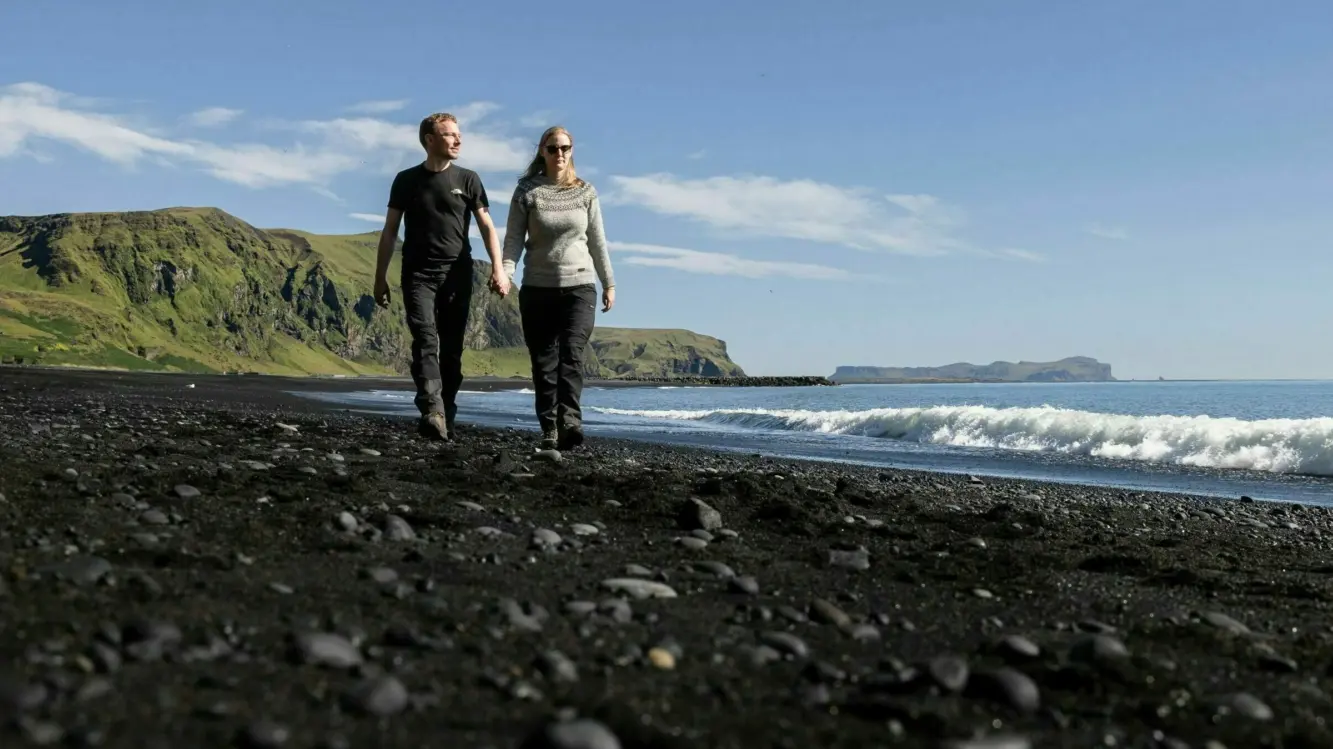 Island strandspaziergang vik timo klingebiel Ein Pärchen welches händchenhaltend an einem schwarzen Strand mit Blick auf Meer und Berge spazieren geht