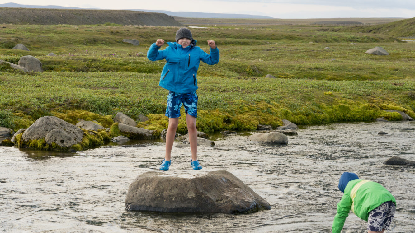 Hochland Kinder spielen am Fluss Kjölur, einer der beiden Jungen steht auf einem Stein in der Mitte des Flusses in blauen Klamotten mit Badehose und ist leicht im Sprung, der andere in grüner Jacke und Badehose greift mit der Hand in den Fluss