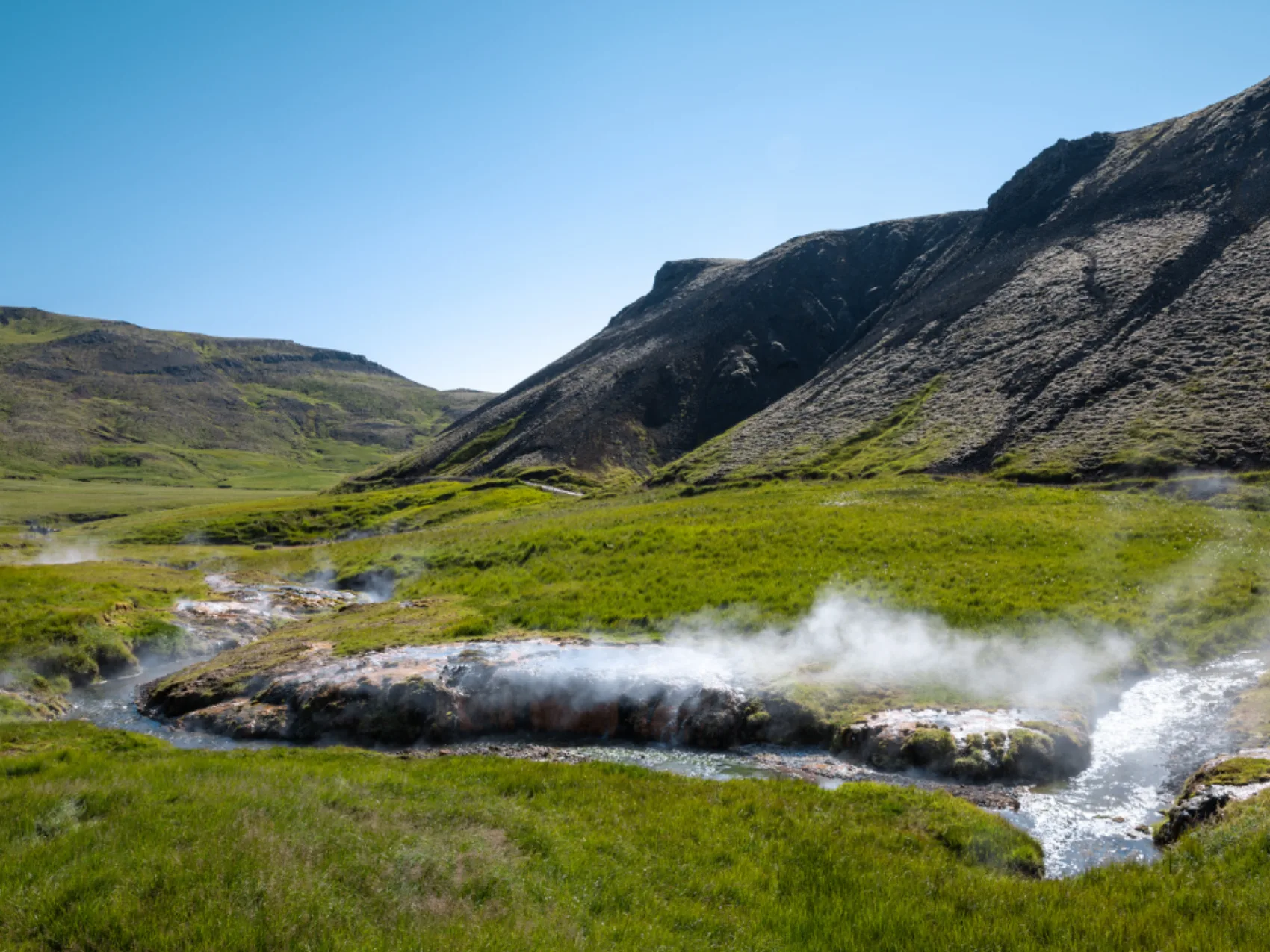 Reykjadalur; 
dampfender Bach schlängelt sich in einem grünen Tal entlang