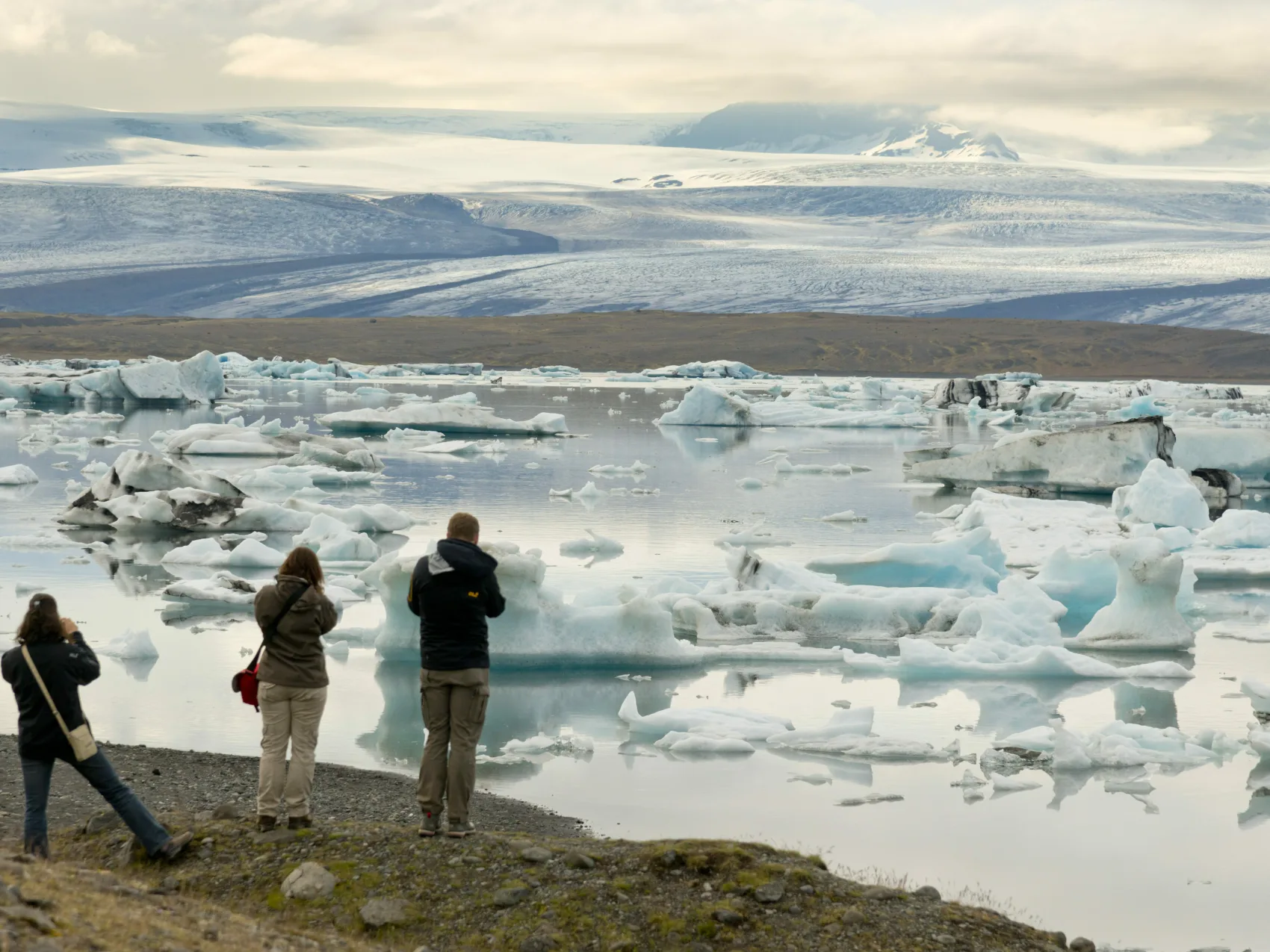 Drei Personen betrachten die Gletscherlagune Jökulsarlon und schießen Fotos. Die Lagune ist voll mit Eisbrocken. Im Hintergrund sieht man den Gletscher Vatnajökull.