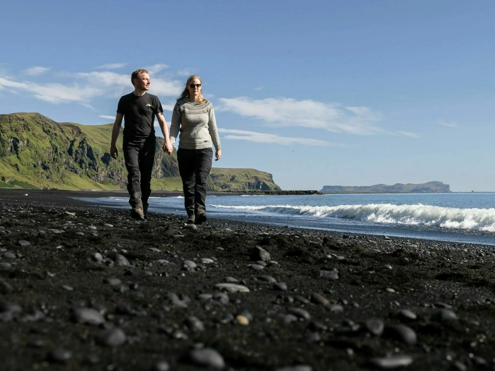 Island Strandspaziergang in Vík, Pärchen läuft Händehaltend am Strand entlang bei Sonnenschein