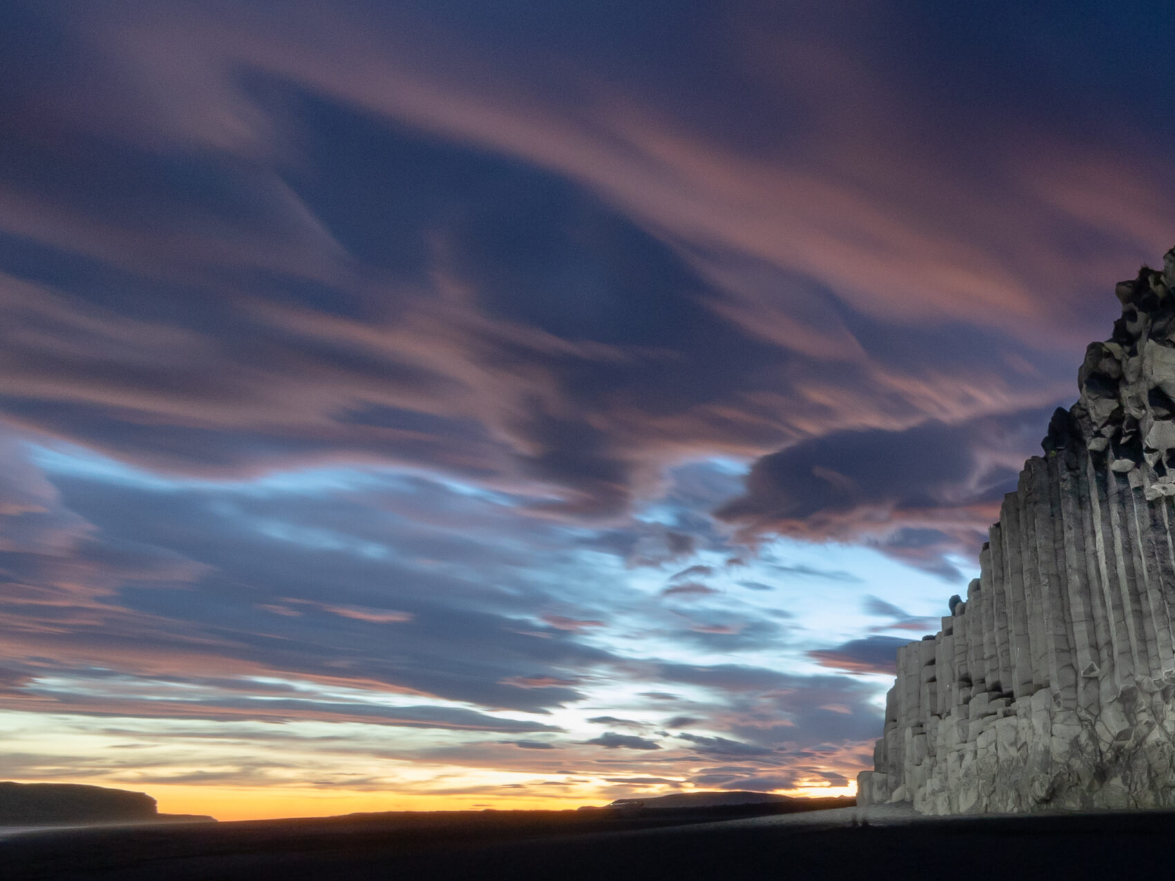 Island Reynisdrangar, Basaltsäulen am rechten Bildrand, schwarzer Sand und lila leuchtende Wolken am Himmel